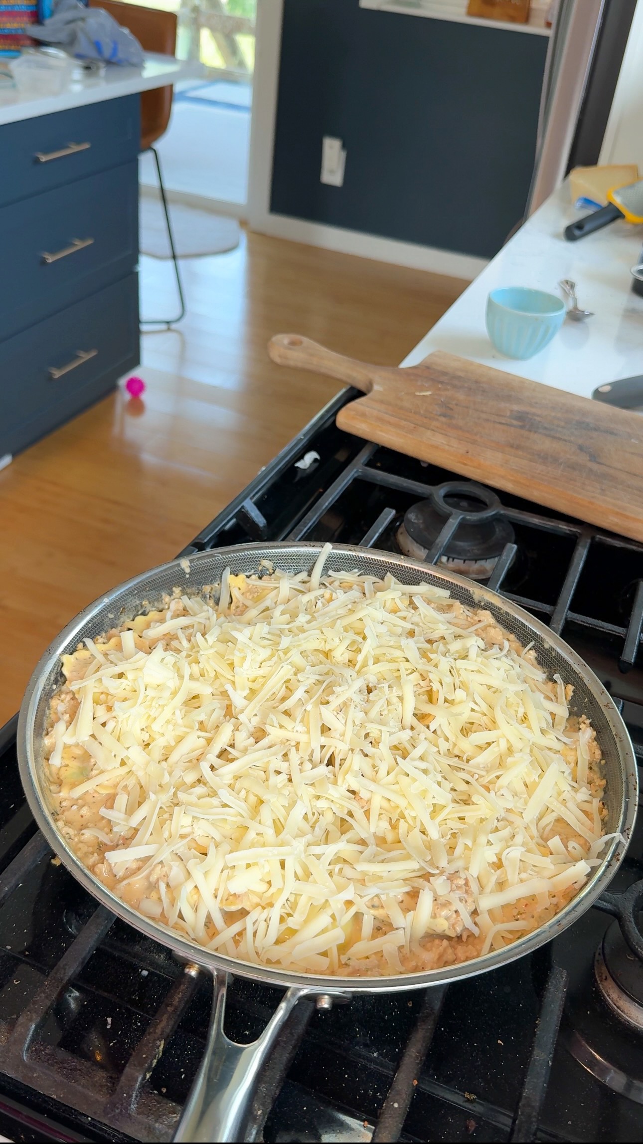 A pan on a stove filled with a dish topped with shredded cheese, ready to be cooked. The kitchen background shows some utensils, a wooden cutting board, and a chair.