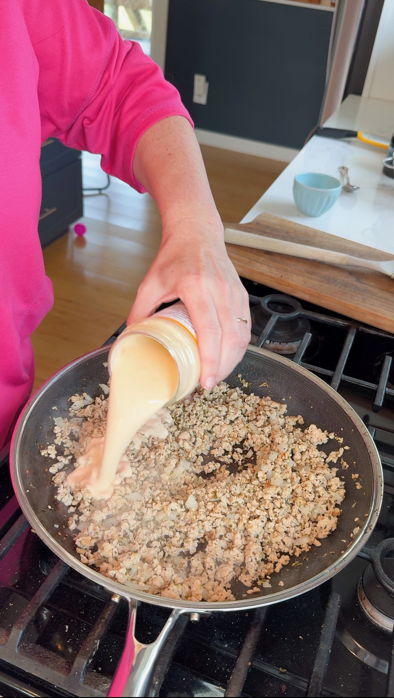 A person pours a creamy sauce from a jar into a skillet filled with browned ground meat on a stove.