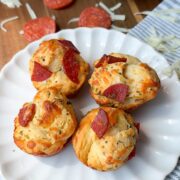 Three pepperoni and cheese muffins sit on a white plate, with shredded cheese, pepperoni slices, and a small blue bowl of marinara sauce in the background. The scene is set on a wooden surface with a striped cloth.