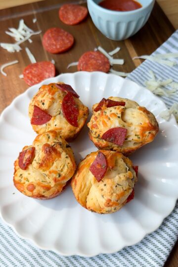 Three pepperoni and cheese muffins sit on a white plate, with shredded cheese, pepperoni slices, and a small blue bowl of marinara sauce in the background. The scene is set on a wooden surface with a striped cloth.