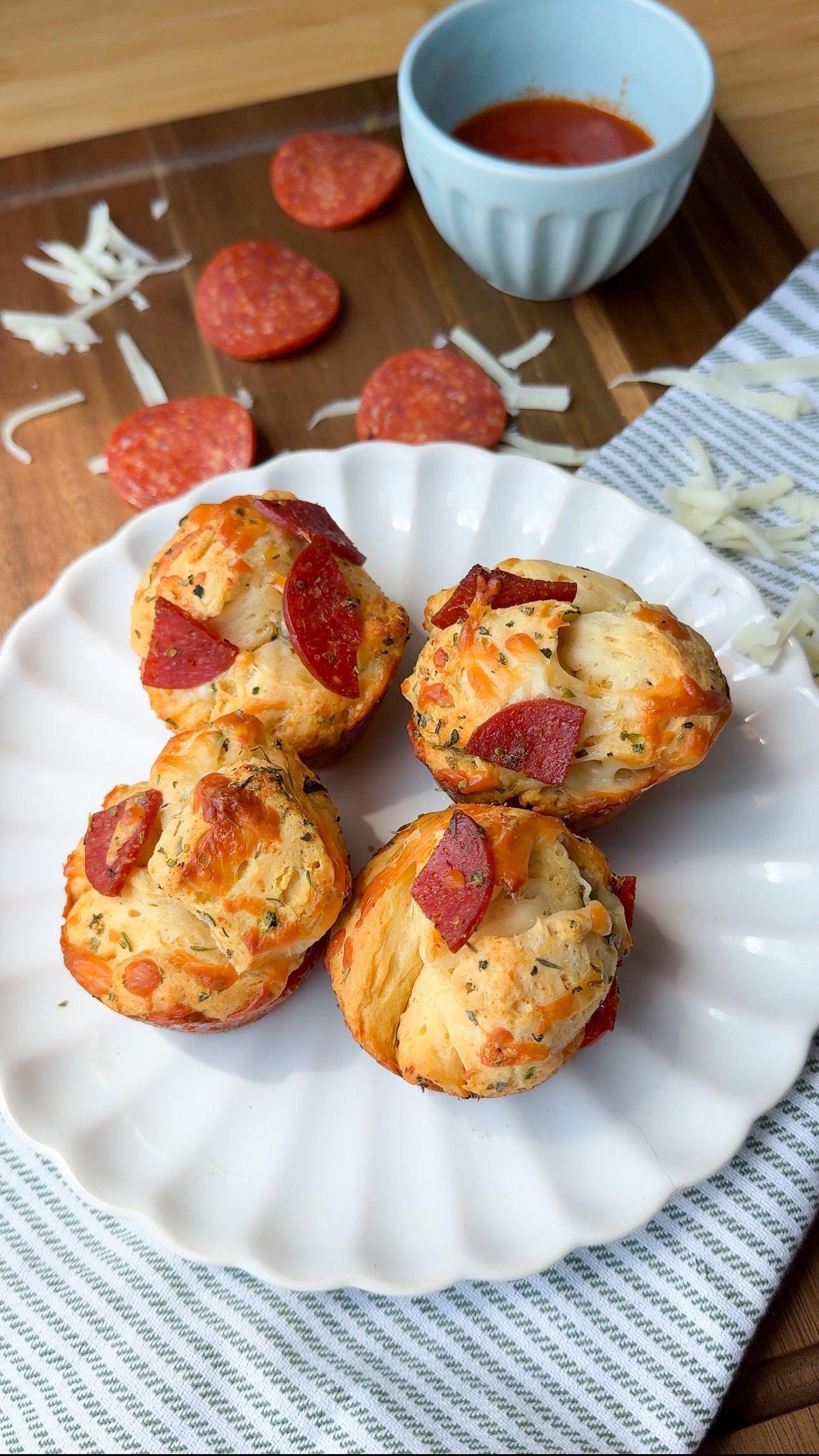 Three pepperoni and cheese muffins sit on a white plate, with shredded cheese, pepperoni slices, and a small blue bowl of marinara sauce in the background. The scene is set on a wooden surface with a striped cloth.