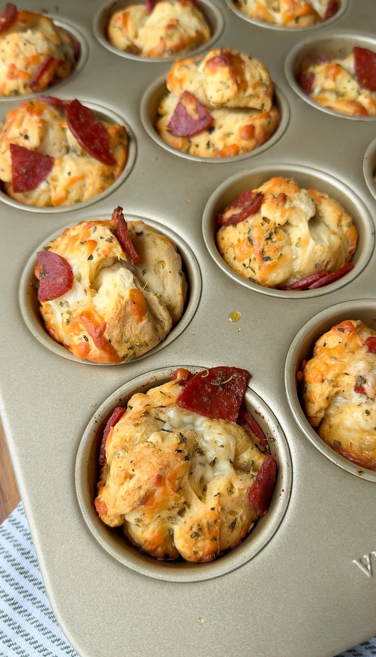 A muffin tin filled with savory muffins containing cheese, herbs, and slices of pepperoni, resting on a striped cloth.