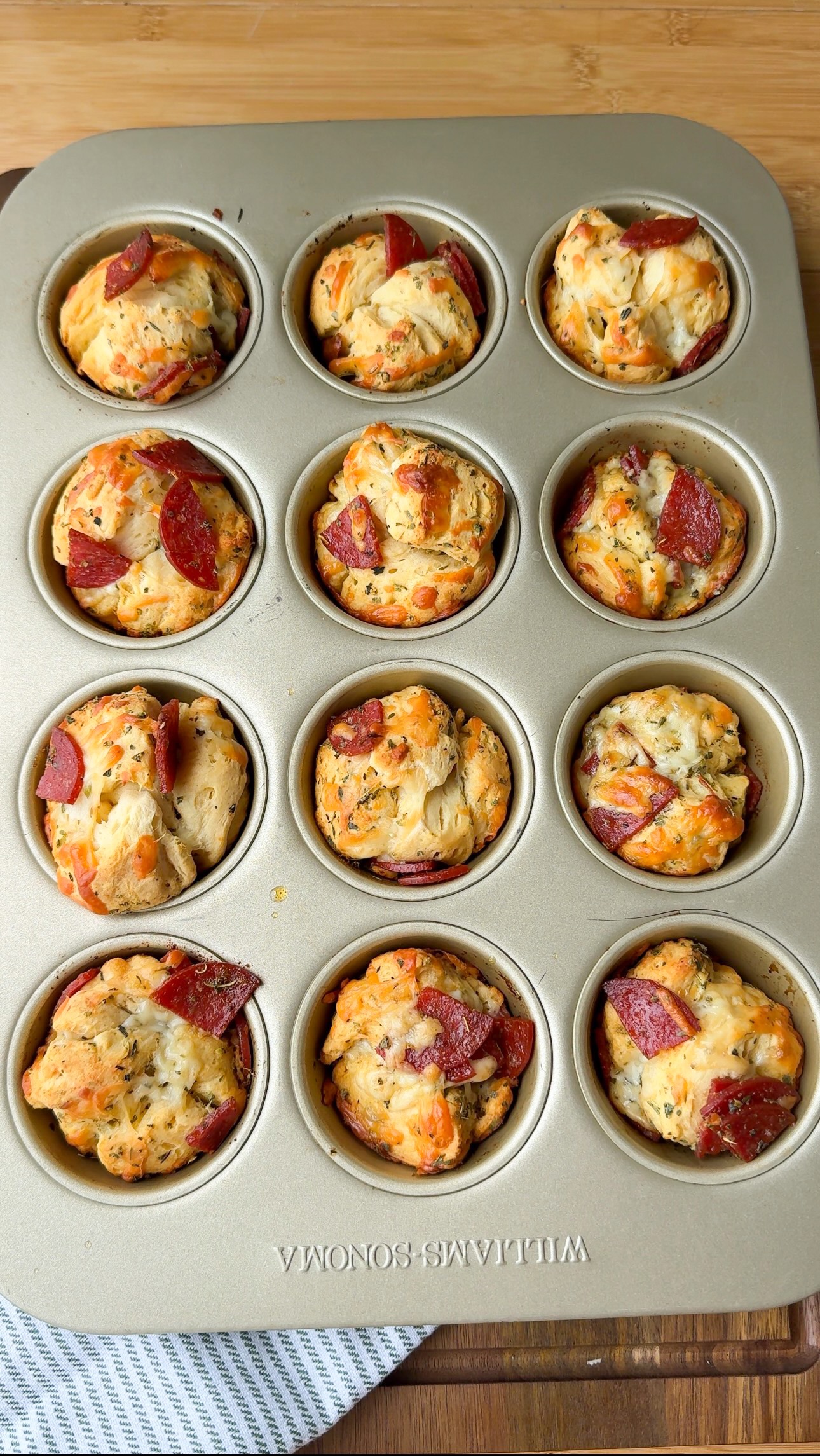 A muffin tin with twelve savory muffins topped with herbs and pepperoni sits on a wooden surface. Some muffins have more pepperoni pieces than others.
