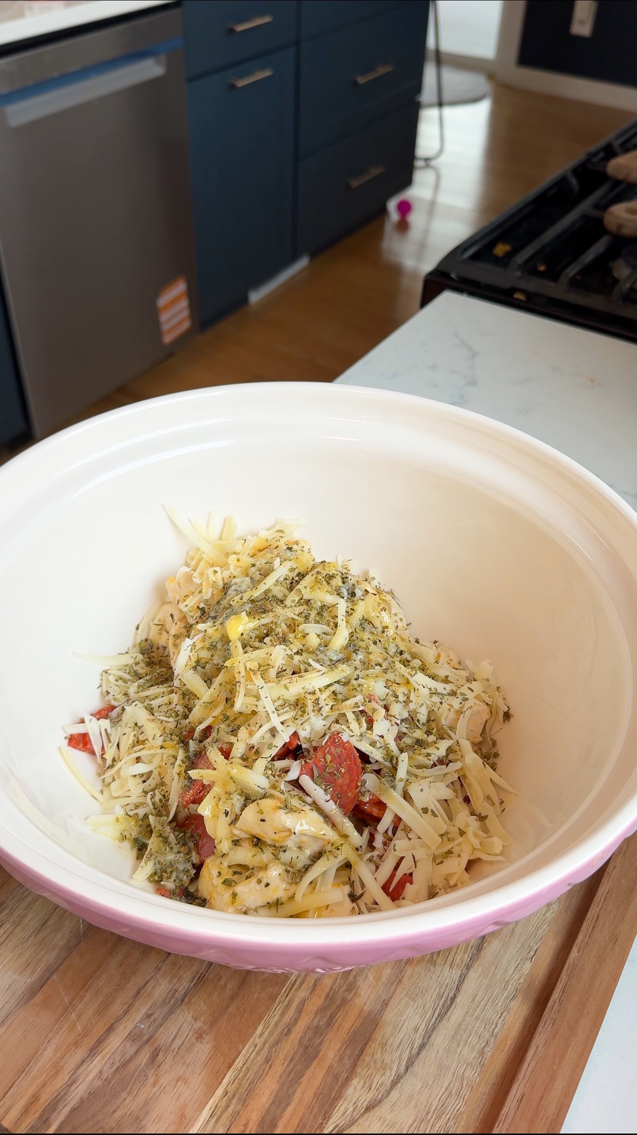A large white and pink bowl filled with shredded cheese, chopped tomatoes, and dried herbs sits on a kitchen counter with blue cabinets in the background.