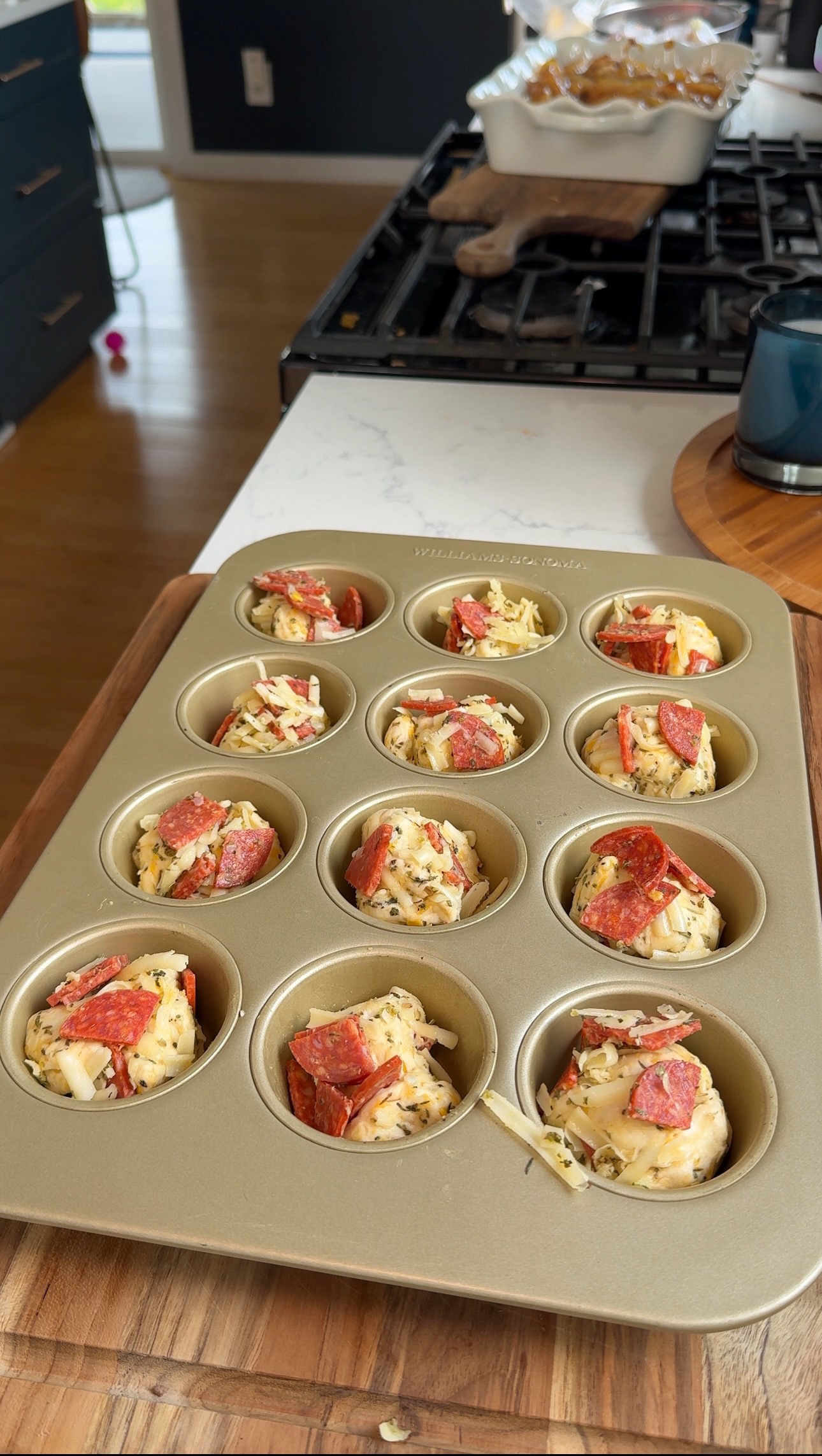 A muffin tin filled with unbaked savory muffins, topped with shredded cheese and pepperoni slices, sits on a kitchen counter near a stove.