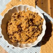 A white bowl filled with chocolate chip cookie dough sits on a striped cloth, surrounded by graham crackers and sprinkled chocolate chips atop a wooden surface.