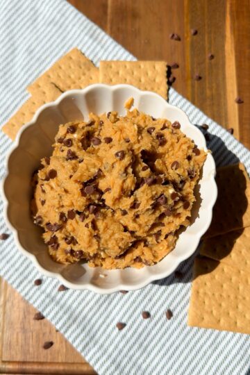 A white bowl filled with chocolate chip cookie dough sits on a striped cloth, surrounded by graham crackers and sprinkled chocolate chips atop a wooden surface.