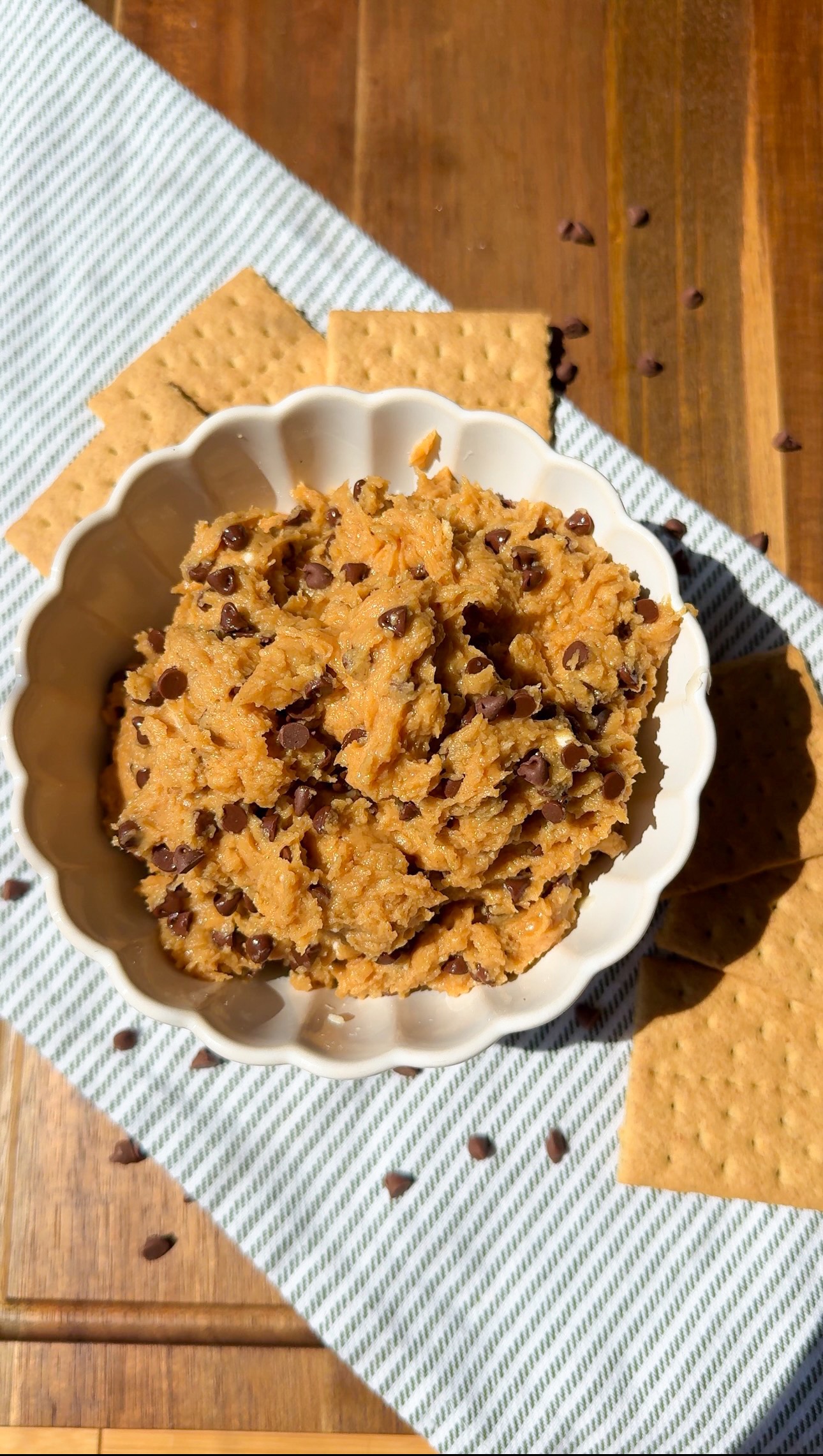 A white bowl filled with chocolate chip cookie dough sits on a striped cloth, surrounded by graham crackers and sprinkled chocolate chips atop a wooden surface.