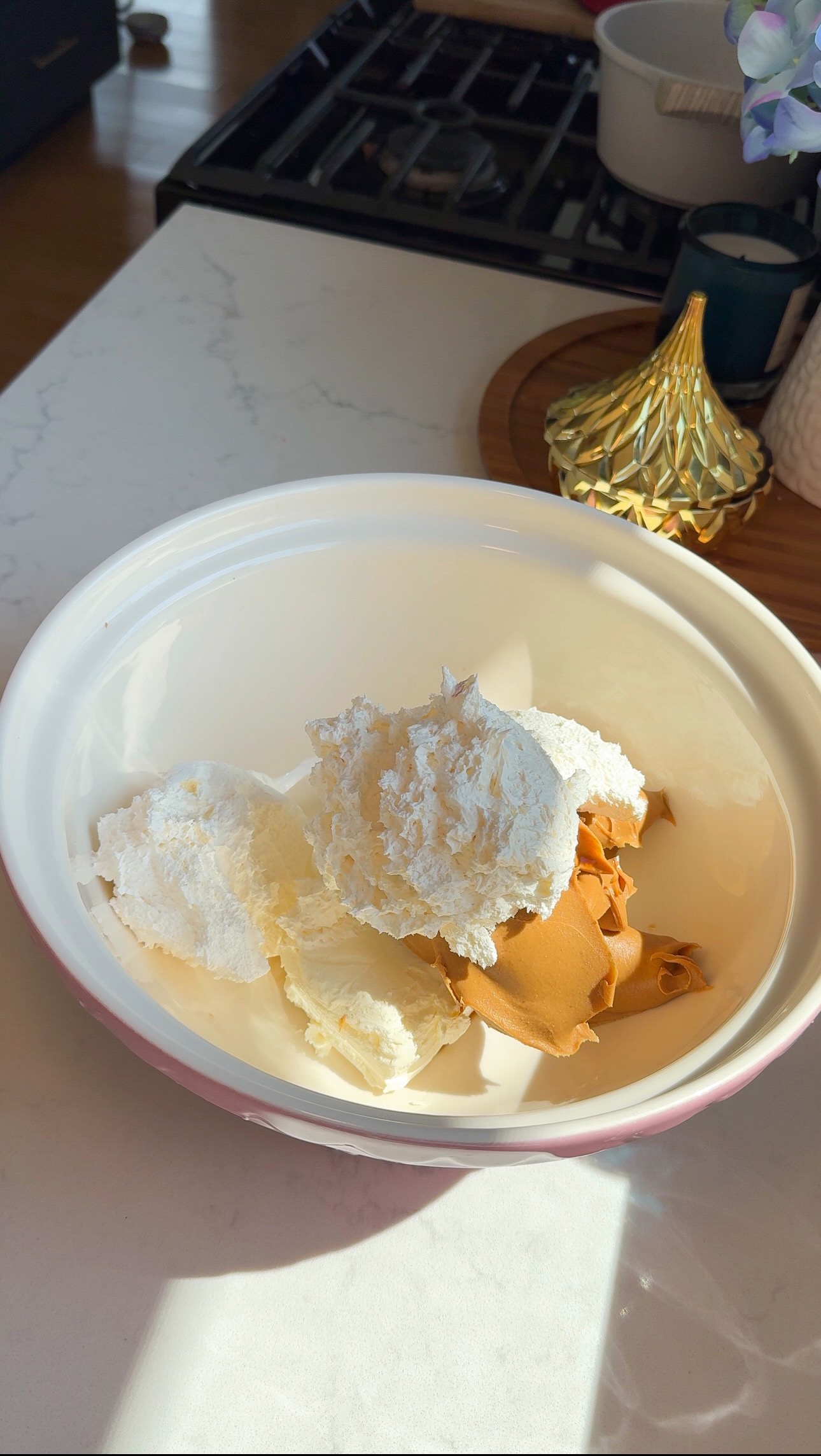 A white bowl containing scoops of whipped topping, cream cheese, and peanut butter sits on a kitchen counter in natural sunlight.