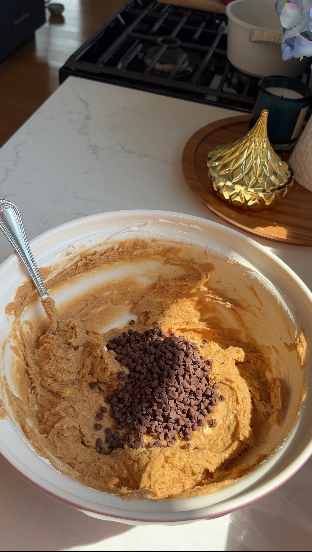 A mixing bowl with cookie dough and chocolate chips sits on a white countertop; a metal spoon rests inside, and sunlight streams onto the scene. A stovetop and decorative items are in the background.