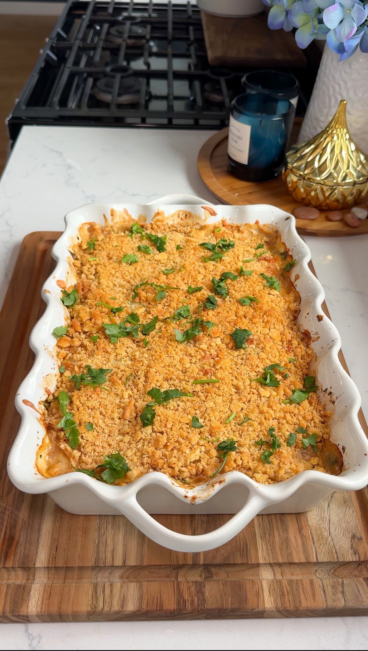 A baked casserole topped with golden breadcrumbs and chopped herbs sits in a white scalloped dish on a wooden board, on a kitchen counter near a stove and decorative items.