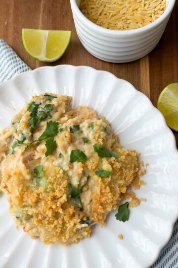 A serving of creamy casserole topped with breadcrumbs and fresh herbs on a white plate, with lime wedges and a bowl of uncooked orzo pasta nearby on a wooden surface.