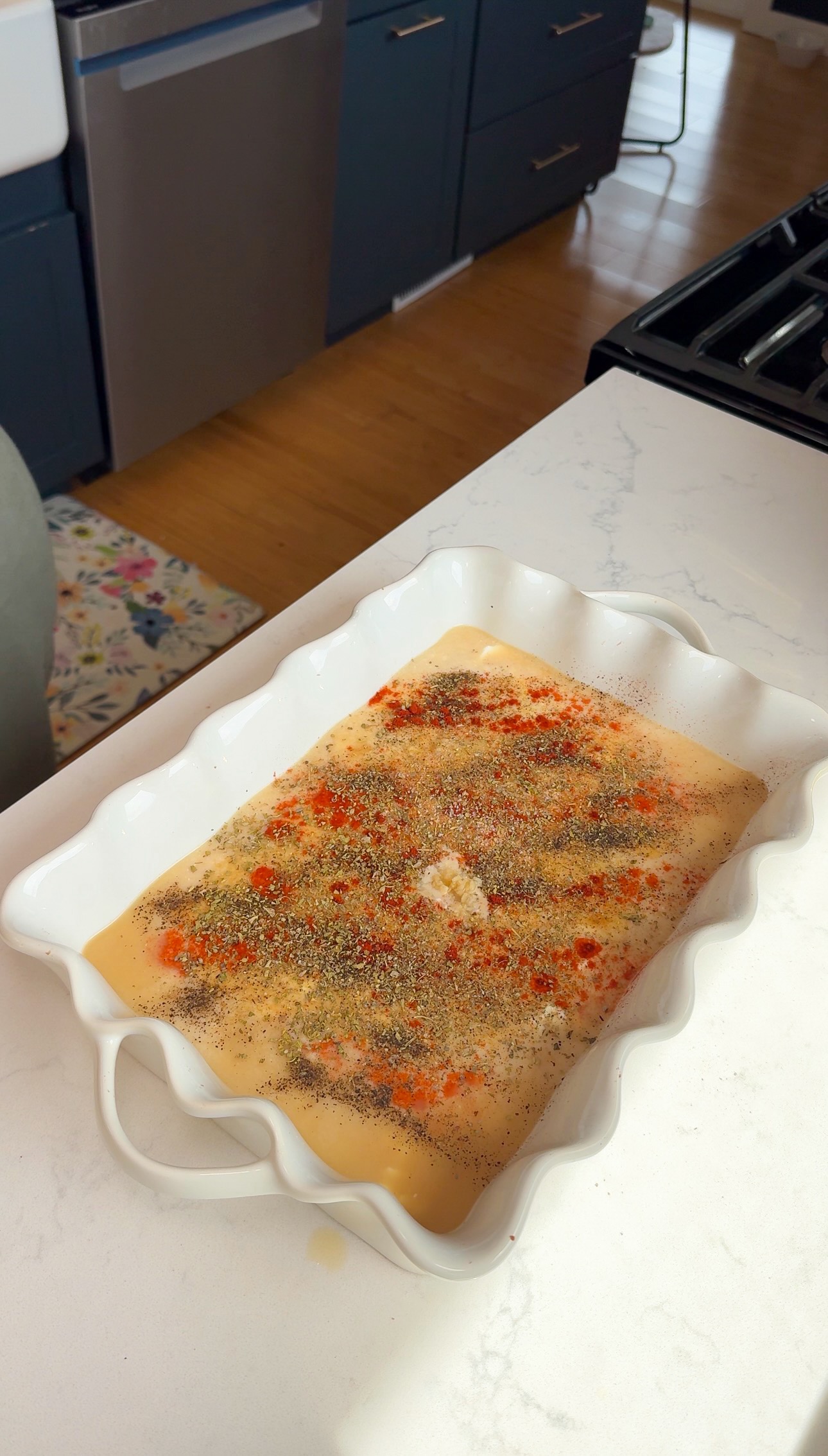 A white baking dish with a liquid mixture, topped with black pepper and red spices, sits on a white countertop in a kitchen.