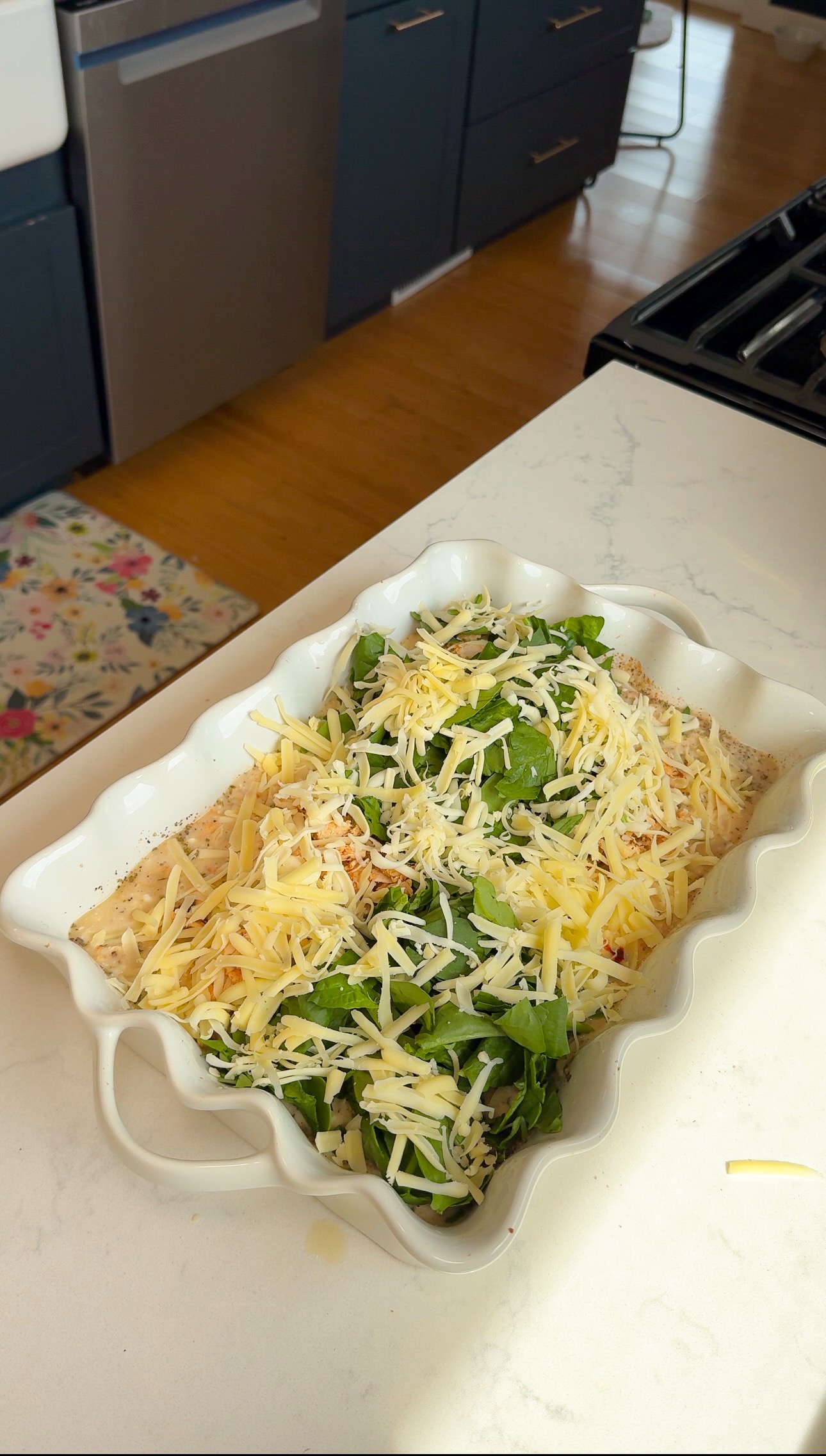 A white baking dish on a kitchen counter contains a casserole topped with shredded cheese and fresh spinach, ready to be baked.