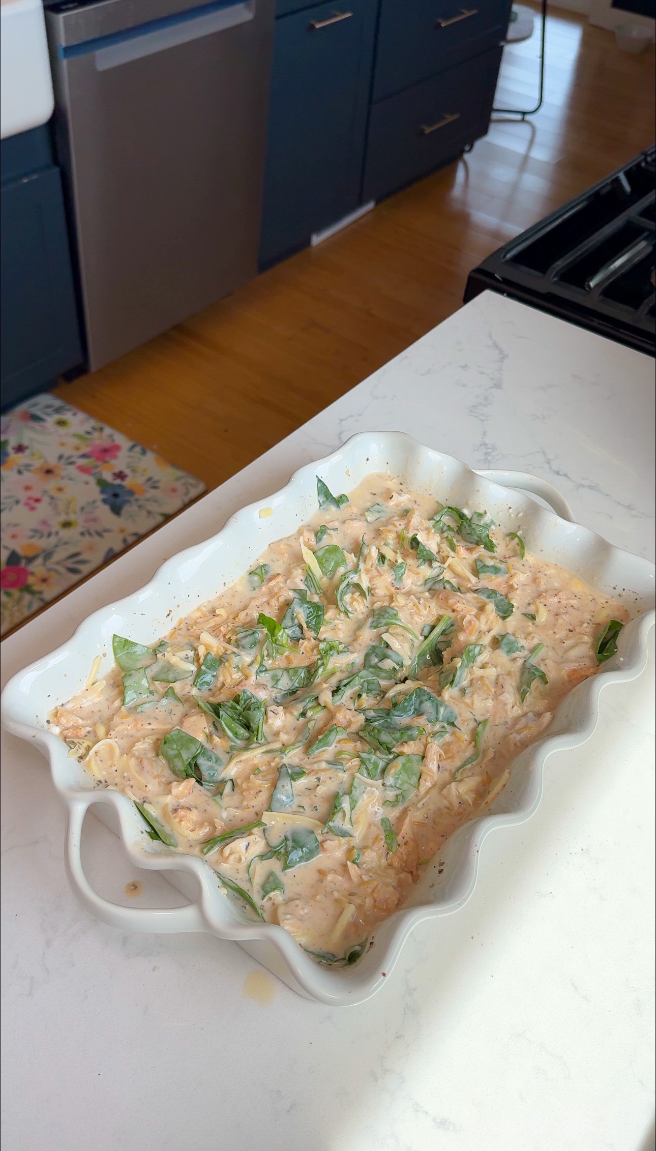 A white ceramic baking dish filled with a creamy pasta bake topped with chopped spinach sits on a white countertop in a modern kitchen.