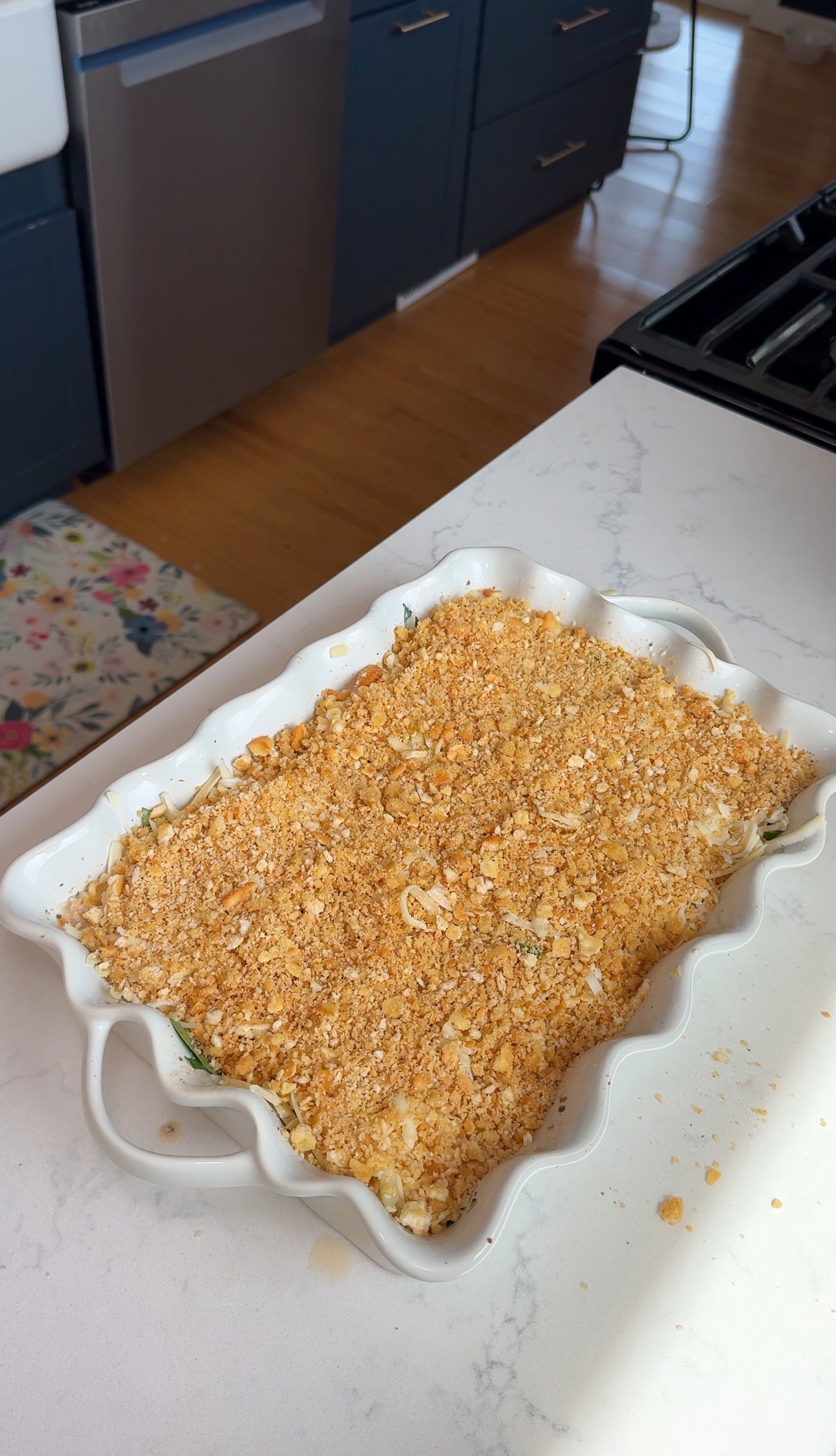 A white baking dish on a marble counter holds a casserole topped with a golden-brown crumb crust, likely breadcrumbs or crushed crackers. Kitchen cabinets, an oven, and a floral rug are visible in the background.