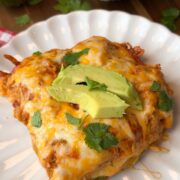 A serving of cheesy enchilada casserole topped with avocado slices and cilantro on a white plate, with tortilla chips and an avocado in the background.