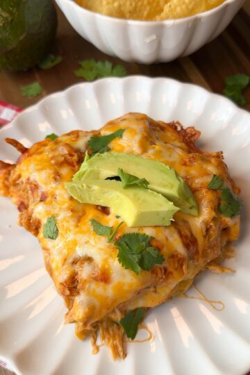 A serving of cheesy enchilada casserole topped with avocado slices and cilantro on a white plate, with tortilla chips and an avocado in the background.