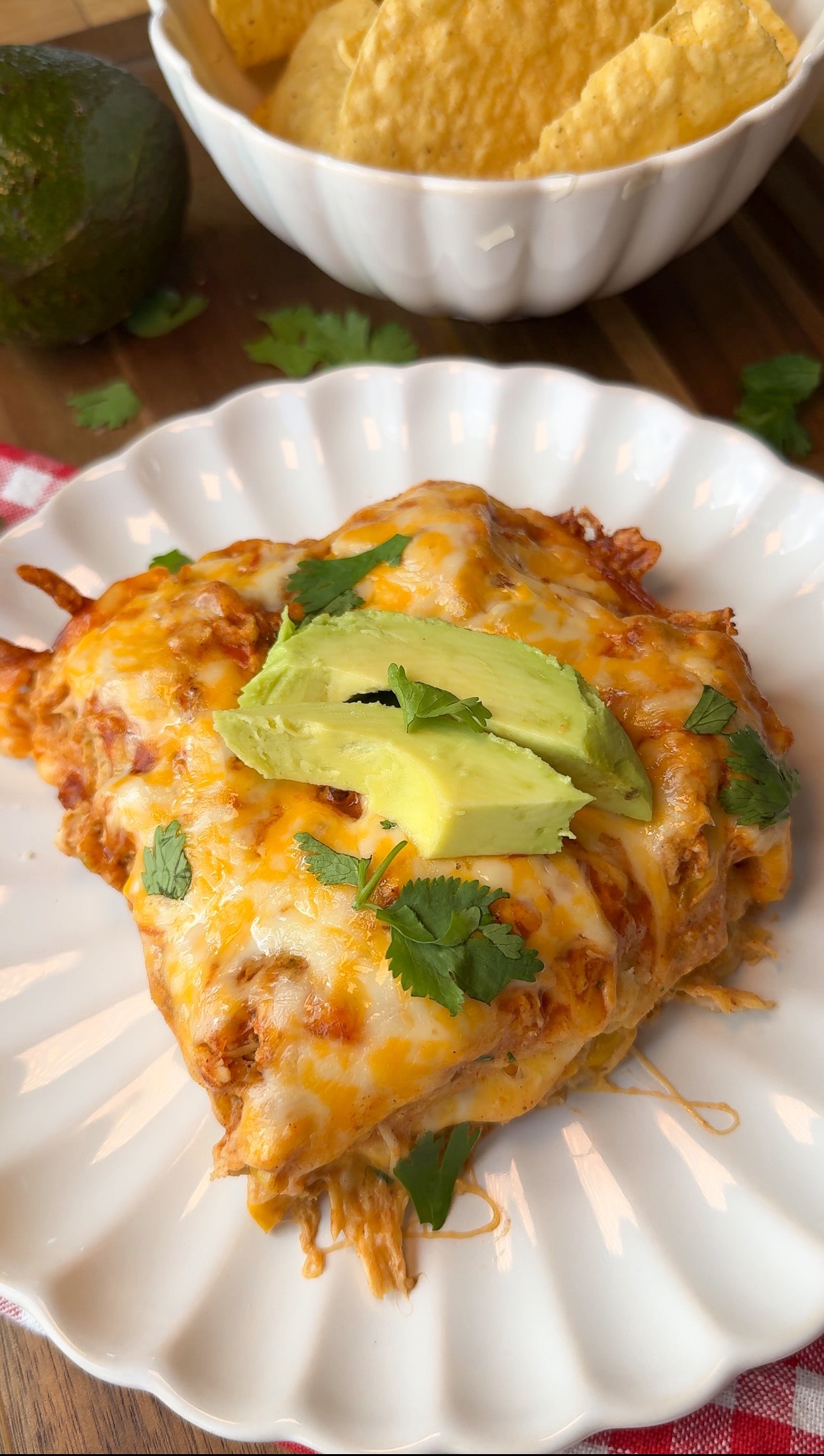 A serving of cheesy enchilada casserole topped with avocado slices and cilantro on a white plate, with tortilla chips and an avocado in the background.