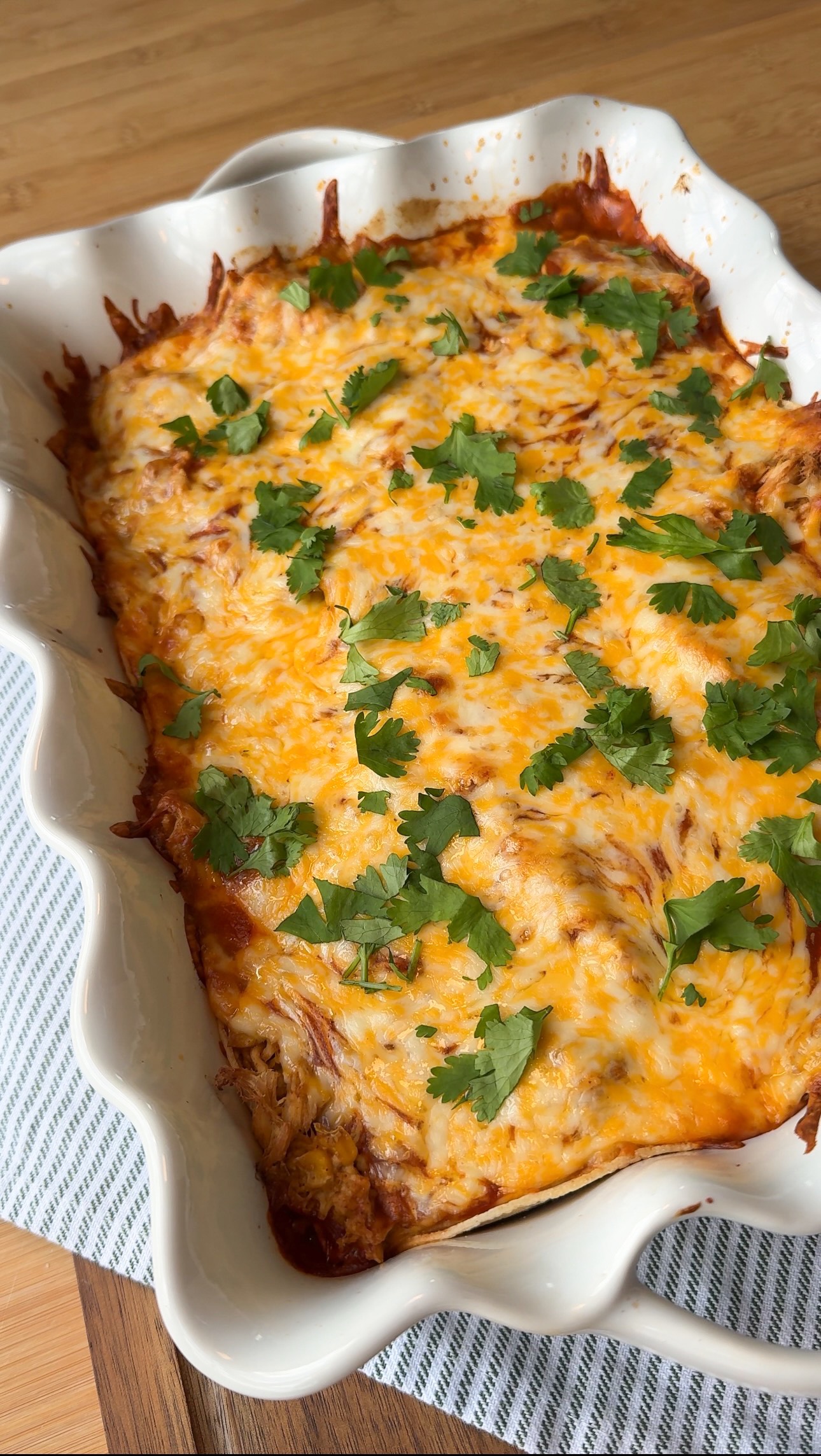 A baked casserole topped with melted cheese and chopped cilantro sits in a white dish, resting on a striped cloth over a wooden surface.