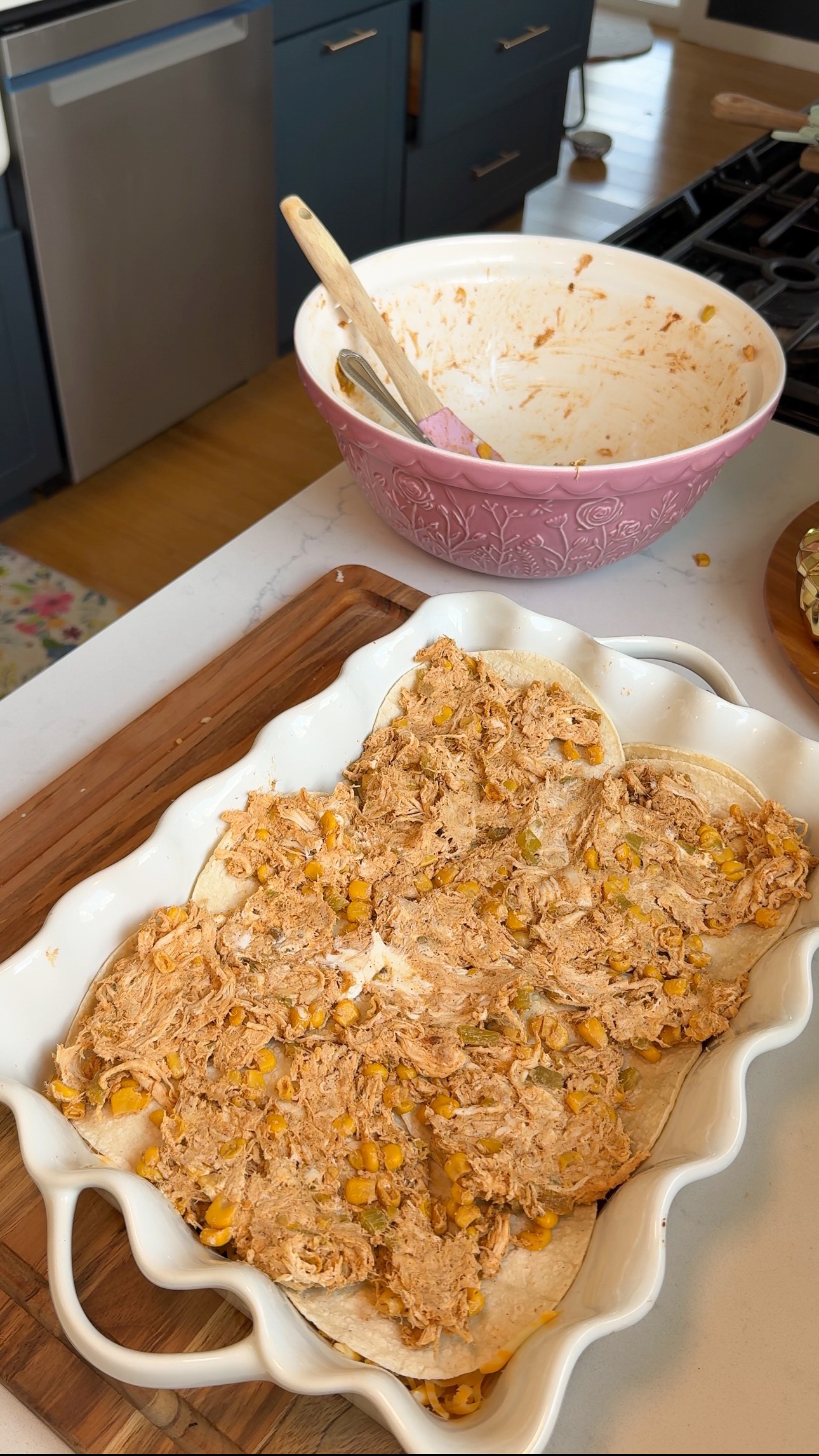 A casserole dish with tortillas topped with shredded chicken, corn, and sauce sits on a counter next to a mixing bowl with a wooden spoon.