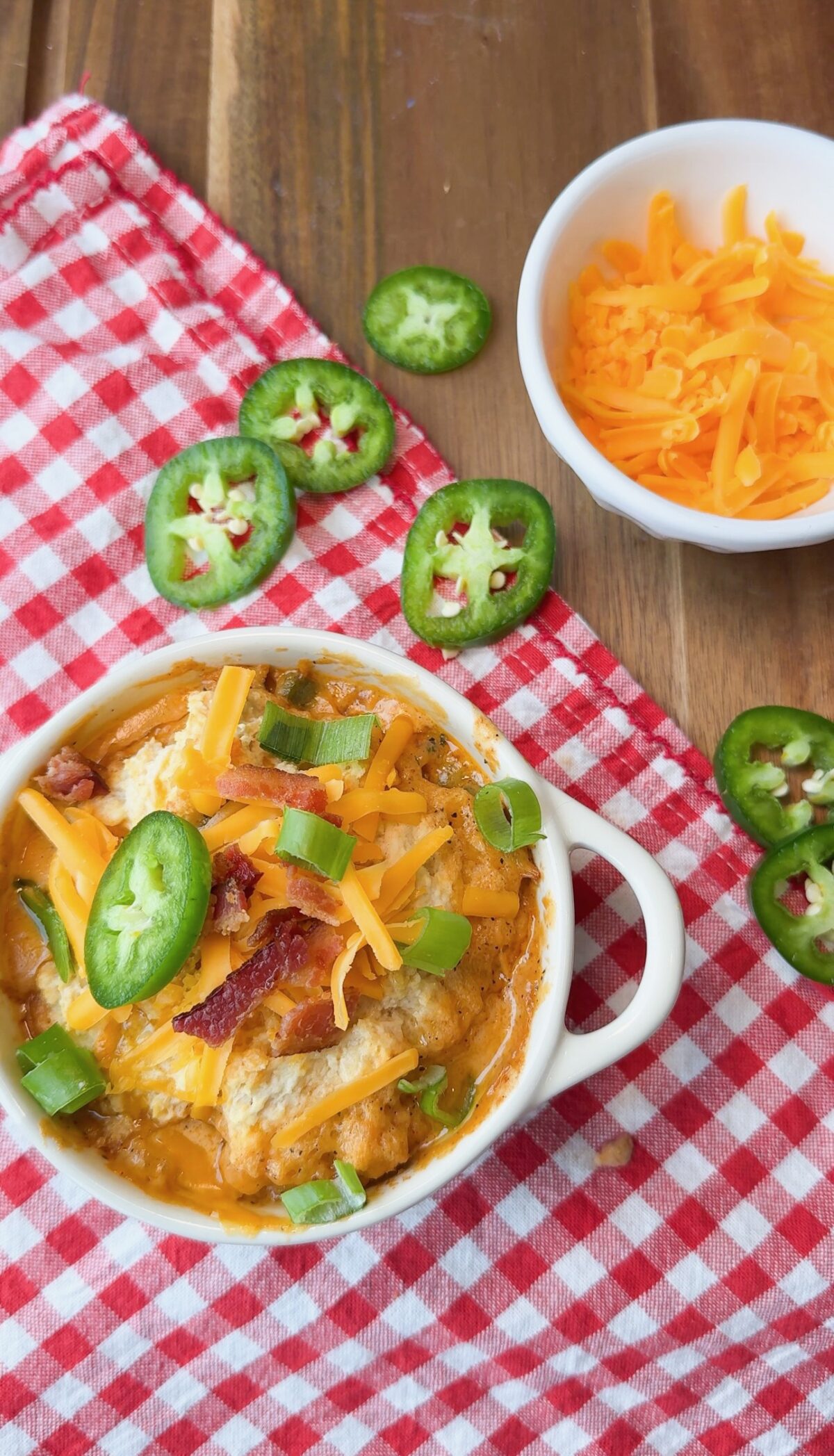 A bowl of cheesy dip topped with bacon, chopped green onions, and jalapeño slices sits on a red checkered cloth. Nearby is a small bowl of shredded cheddar cheese and a few more jalapeño slices.