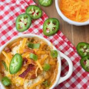 A bowl of cheesy dip topped with bacon, chopped green onions, and jalapeño slices sits on a red checkered cloth. Nearby is a small bowl of shredded cheddar cheese and a few more jalapeño slices.