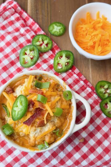 A bowl of cheesy dip topped with bacon, chopped green onions, and jalapeño slices sits on a red checkered cloth. Nearby is a small bowl of shredded cheddar cheese and a few more jalapeño slices.