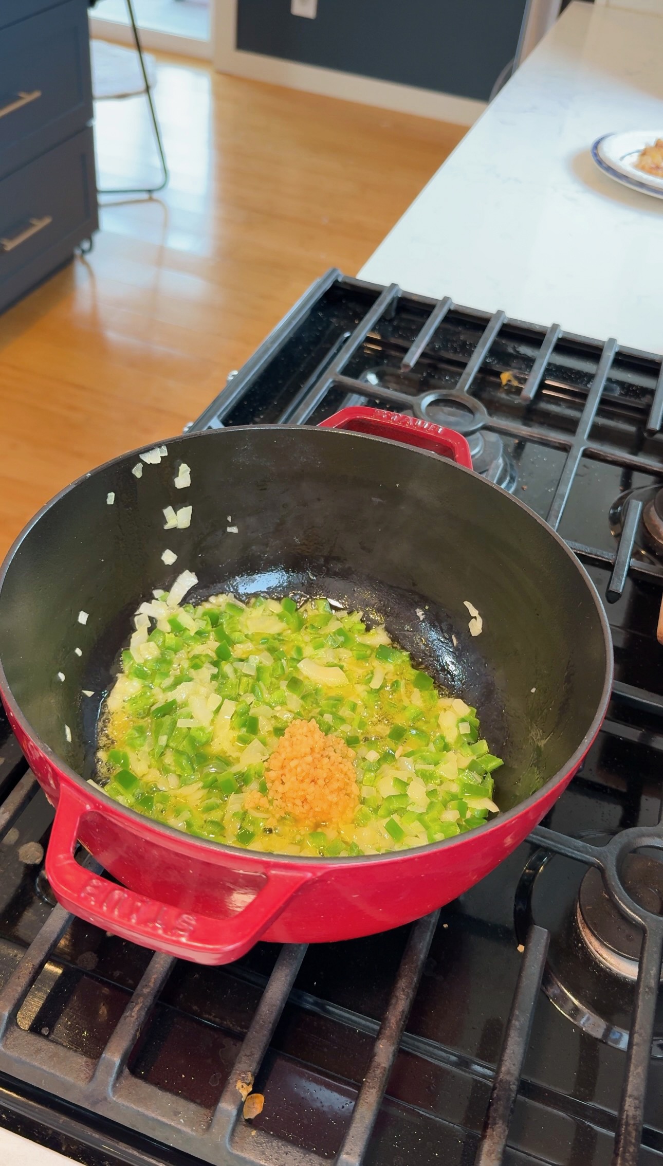 Chopped onions, green peppers, and minced garlic sauté in oil inside a red Dutch oven on a stovetop. The mixture is starting to soften and brown.