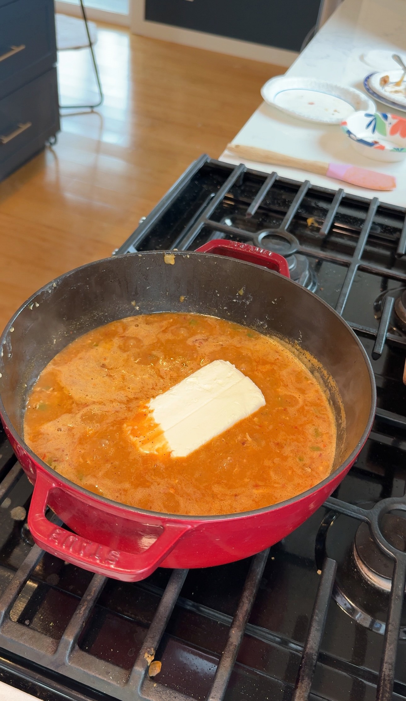 A red pot on a stove contains an orange-brown simmering sauce with a block of cream cheese melting in the center. Nearby, kitchen items rest on a white counter.