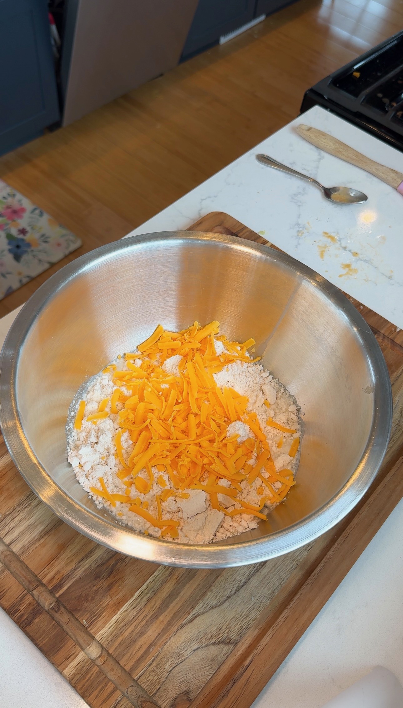 A metal mixing bowl with shredded cheddar cheese and flour sits on a wooden cutting board, next to a spoon and countertop in a kitchen.