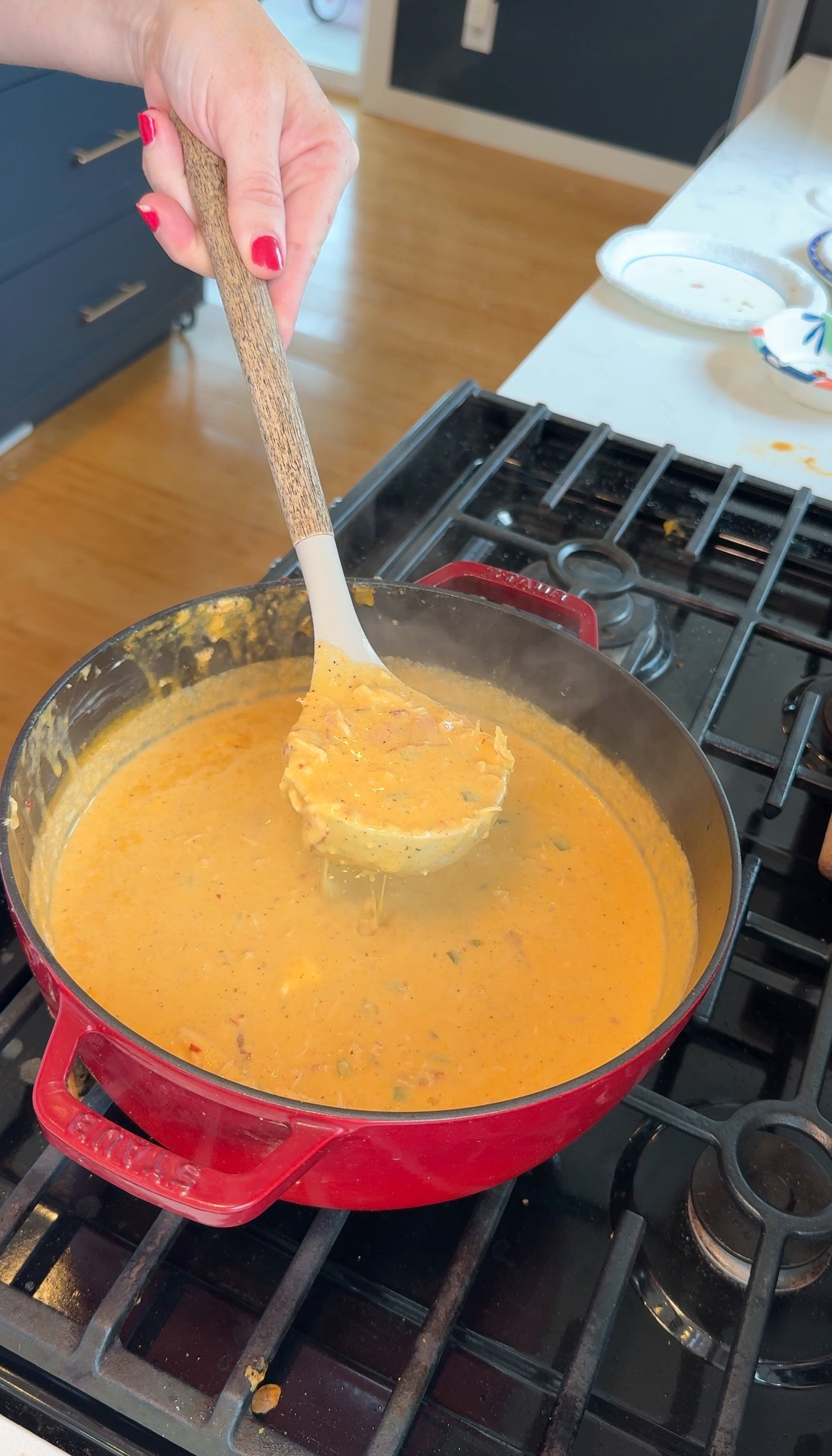 A hand holds a ladle above a red pot of creamy orange soup or stew cooking on a stove, with wooden flooring and kitchen counters visible in the background.