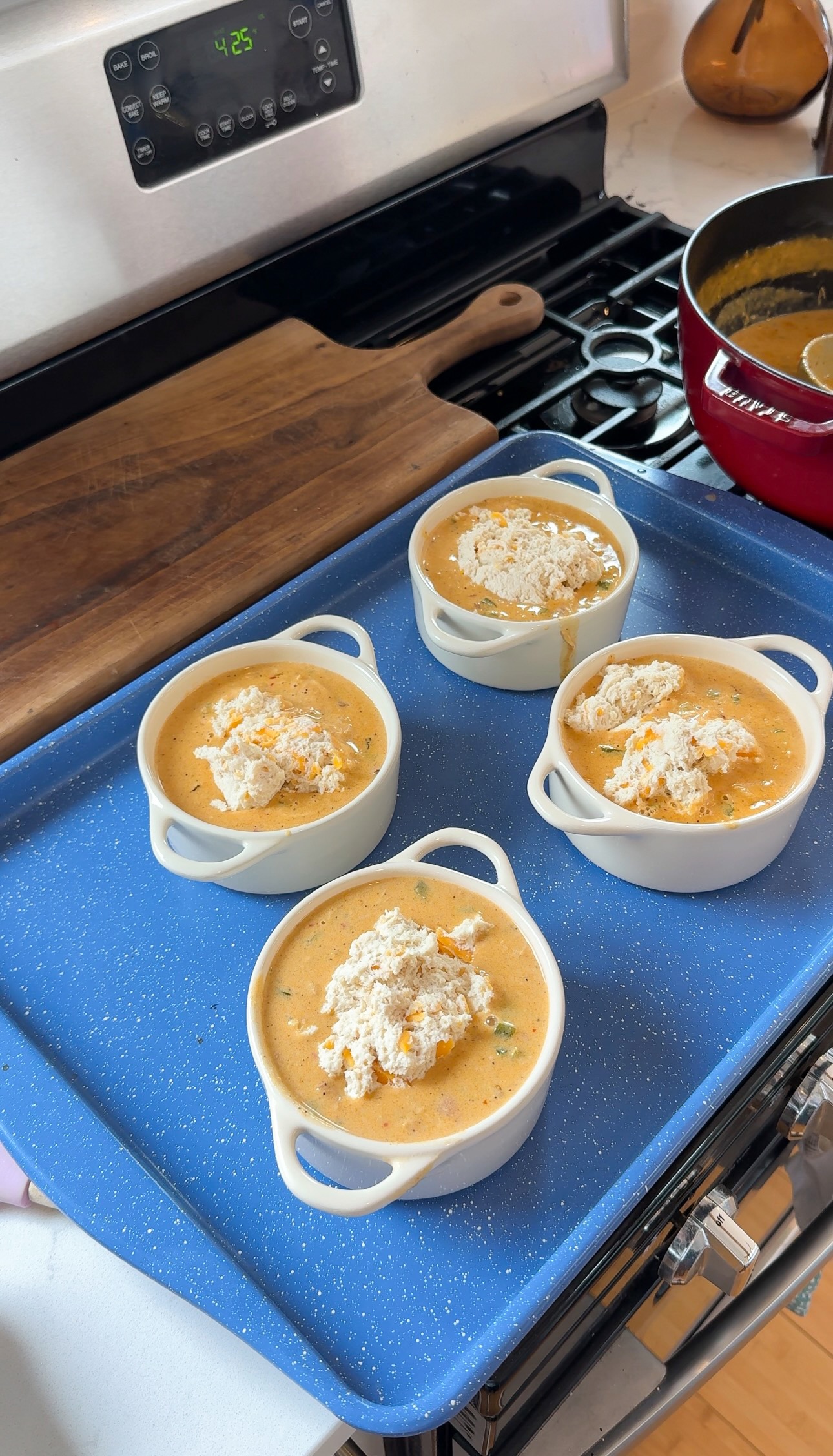 Four small white ramekins filled with a creamy orange soup, topped with biscuit dough, sit on a blue tray on a stove next to a pot and wooden cutting board.