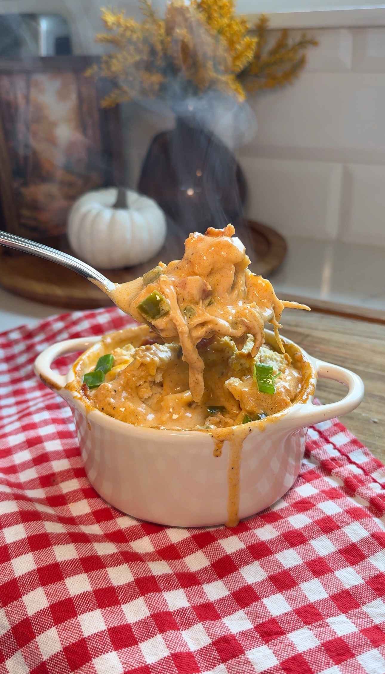 A steaming cheesy casserole with peppers is lifted by a fork from a small white dish, placed on a red and white checkered cloth. A decorative white pumpkin sits in the background.