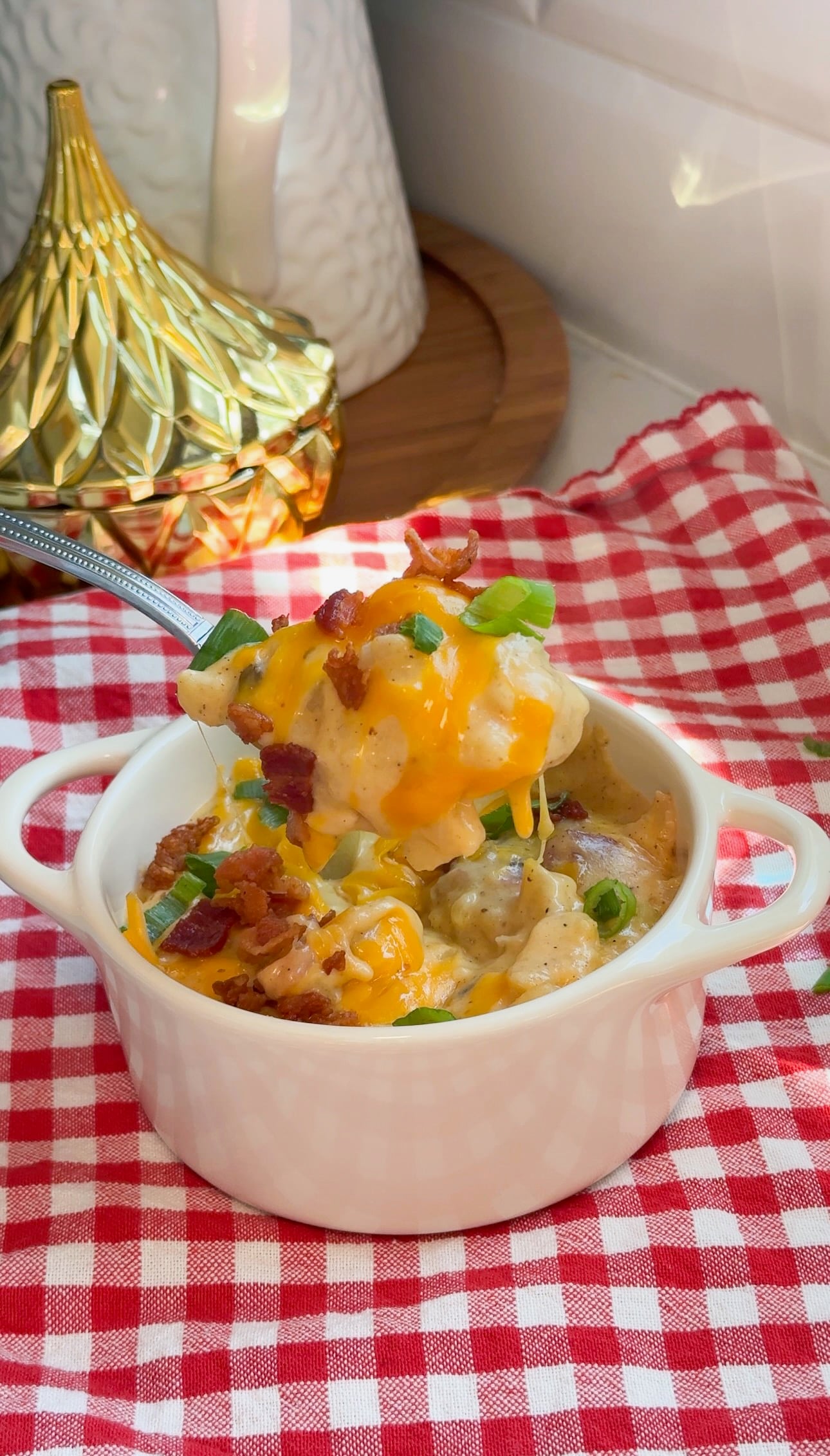 A white ramekin with cheesy potato casserole topped with bacon and green onions, on a red checkered cloth. A spoon lifts a portion, and a gold decorative object is in the background.