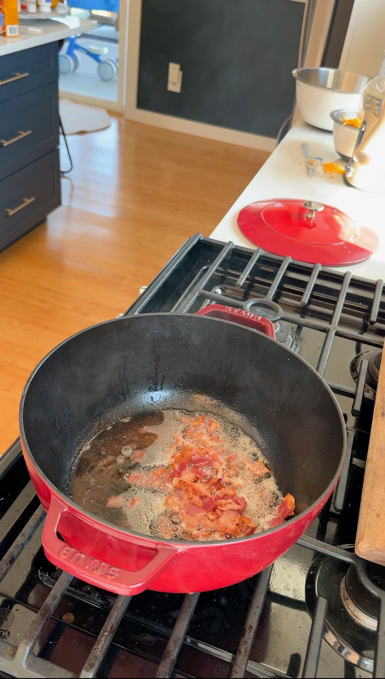 Red dutch oven with sizzling bacon pieces cooking on a stovetop. Nearby, a red lid, wooden spoon, and mixing bowl with cracked eggs and whisk are visible on the white countertop. Kitchen scene with bright lighting.