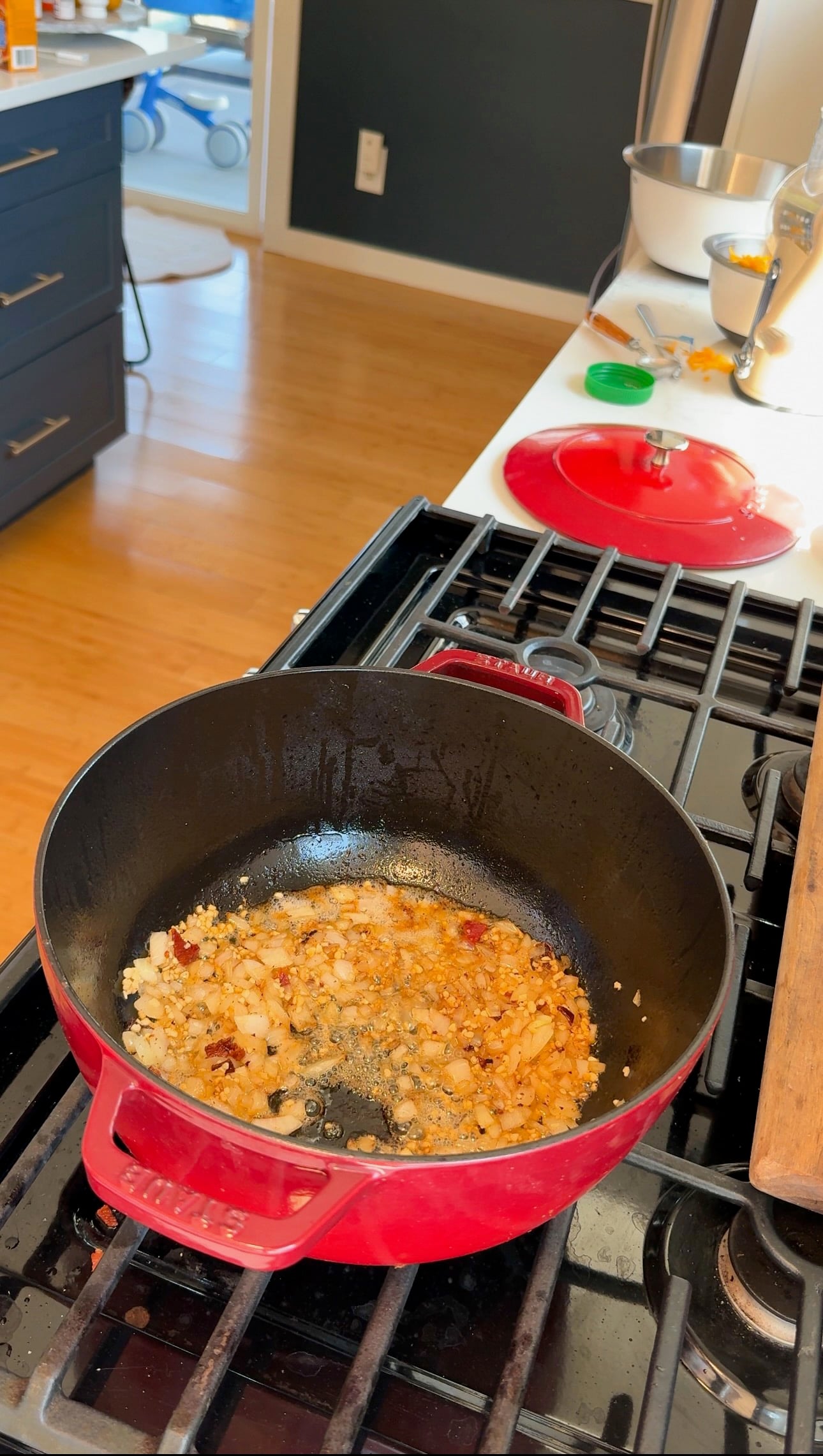 Chopped onions and garlic are sautéing in a red Dutch oven on a stove. Kitchen counters, utensils, and a mixing bowl are visible in the background.