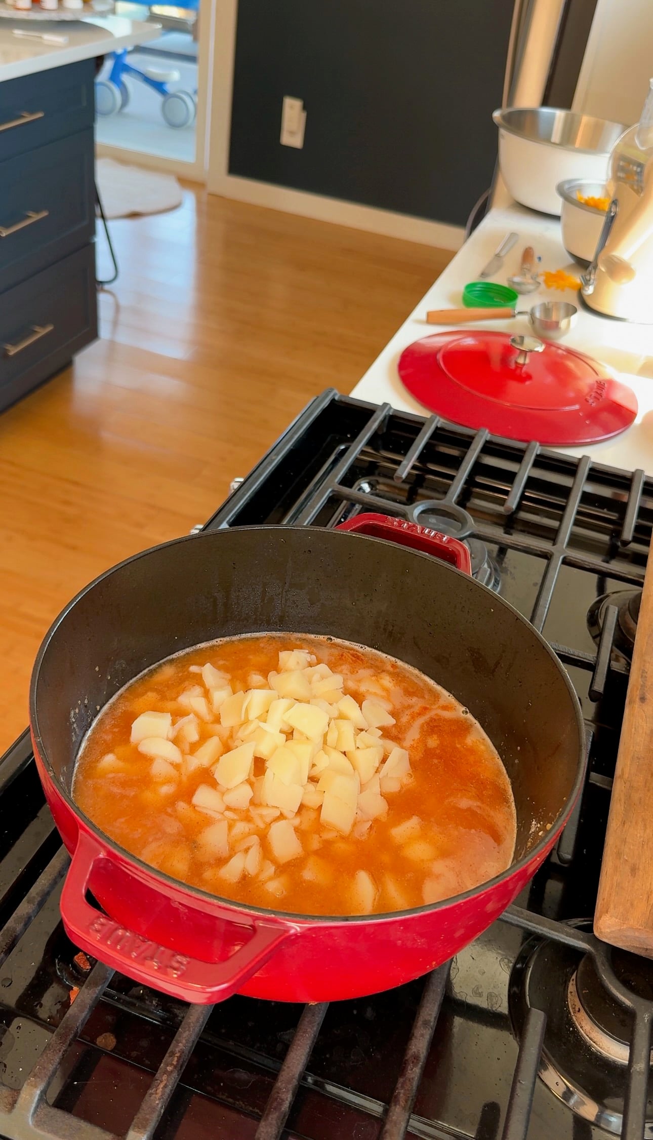 Red pot on a stove filled with soup or stew, topped with diced potatoes. Kitchen utensils and ingredients are on the counter nearby. Sunlight streams in through a window, illuminating the wooden floor and modern kitchen.
