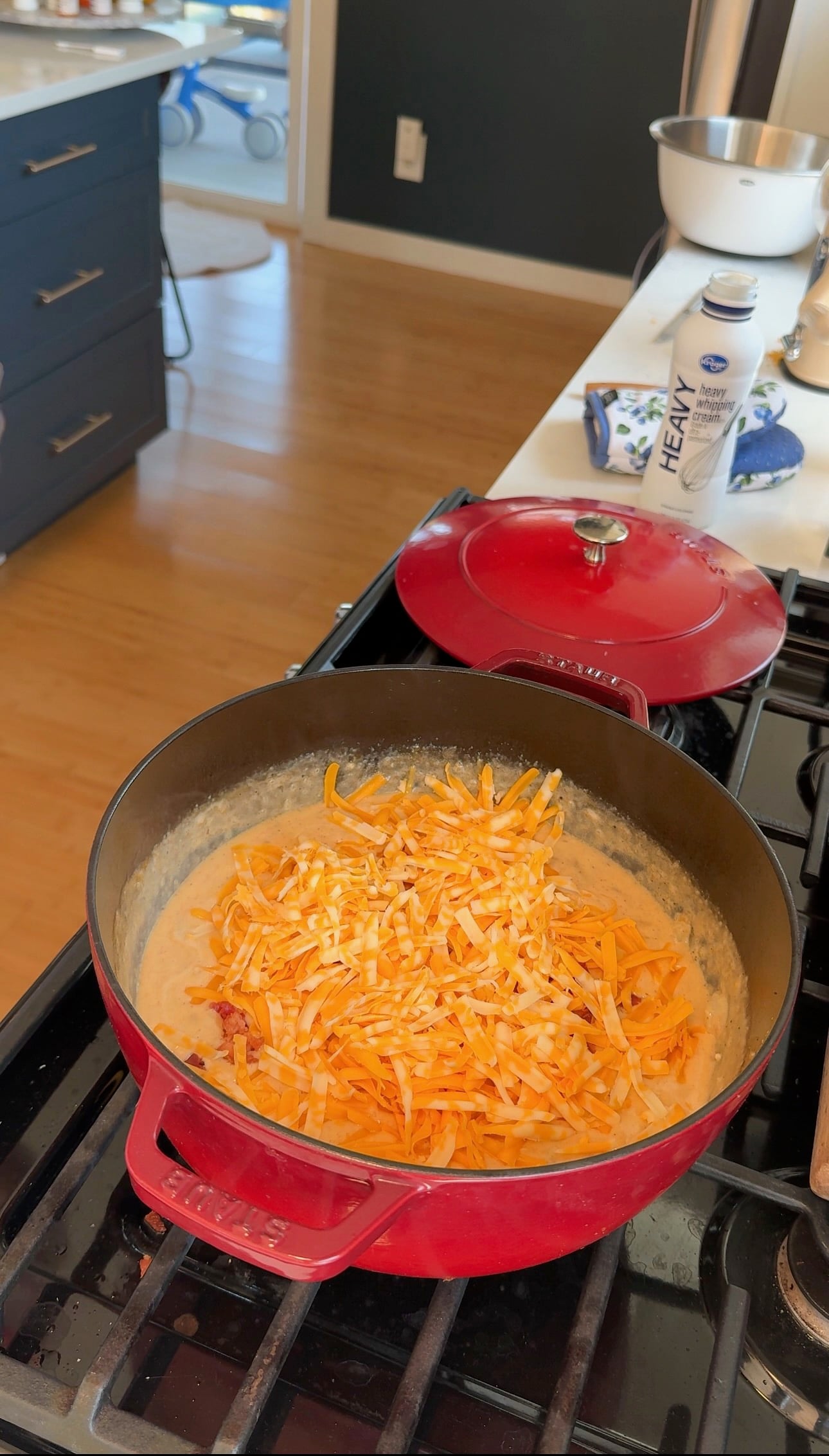 A red pot on a stove contains a creamy mixture topped with shredded cheddar and Colby jack cheese. A red lid sits nearby, and a carton of heavy cream is on the white countertop in the background.