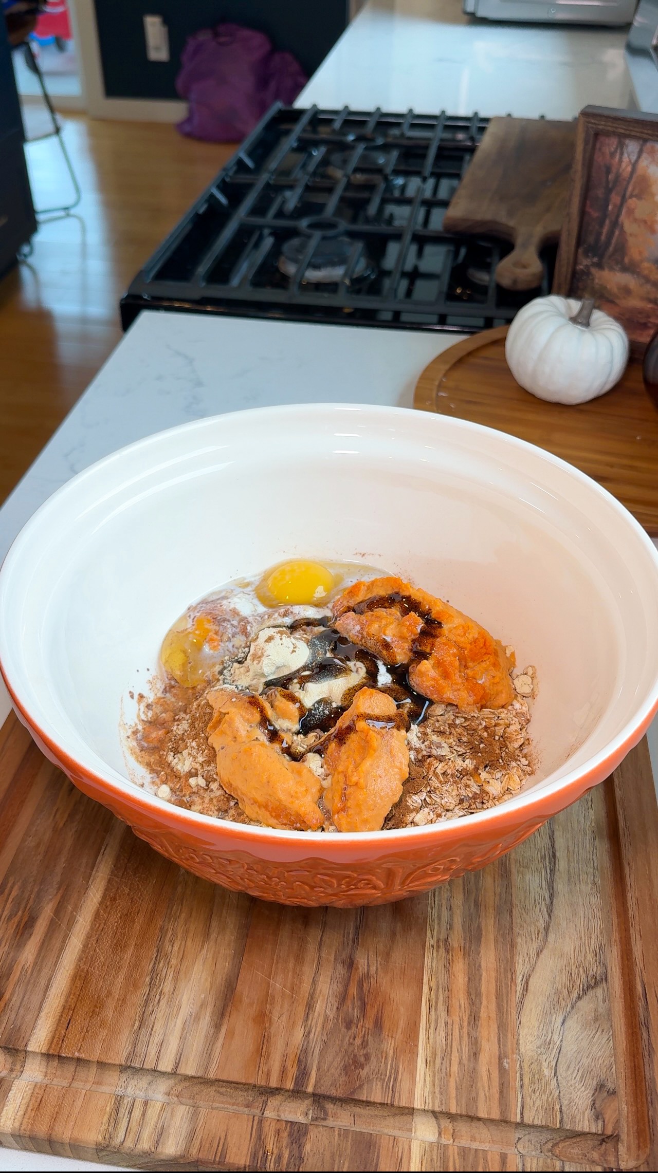 A large bowl with eggs, pumpkin puree, spices, oats, and syrup sits on a wooden board in a kitchen, ready to be mixed for baking.