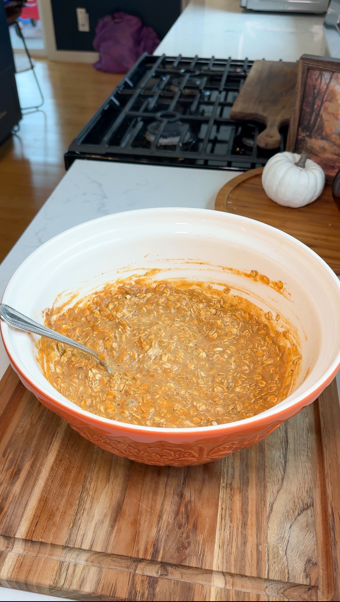 A mixing bowl with oatmeal and pumpkin mixture sits on a wooden cutting board, with a spoon inside. A stove, cutting board, and white decorative pumpkin are in the background.
