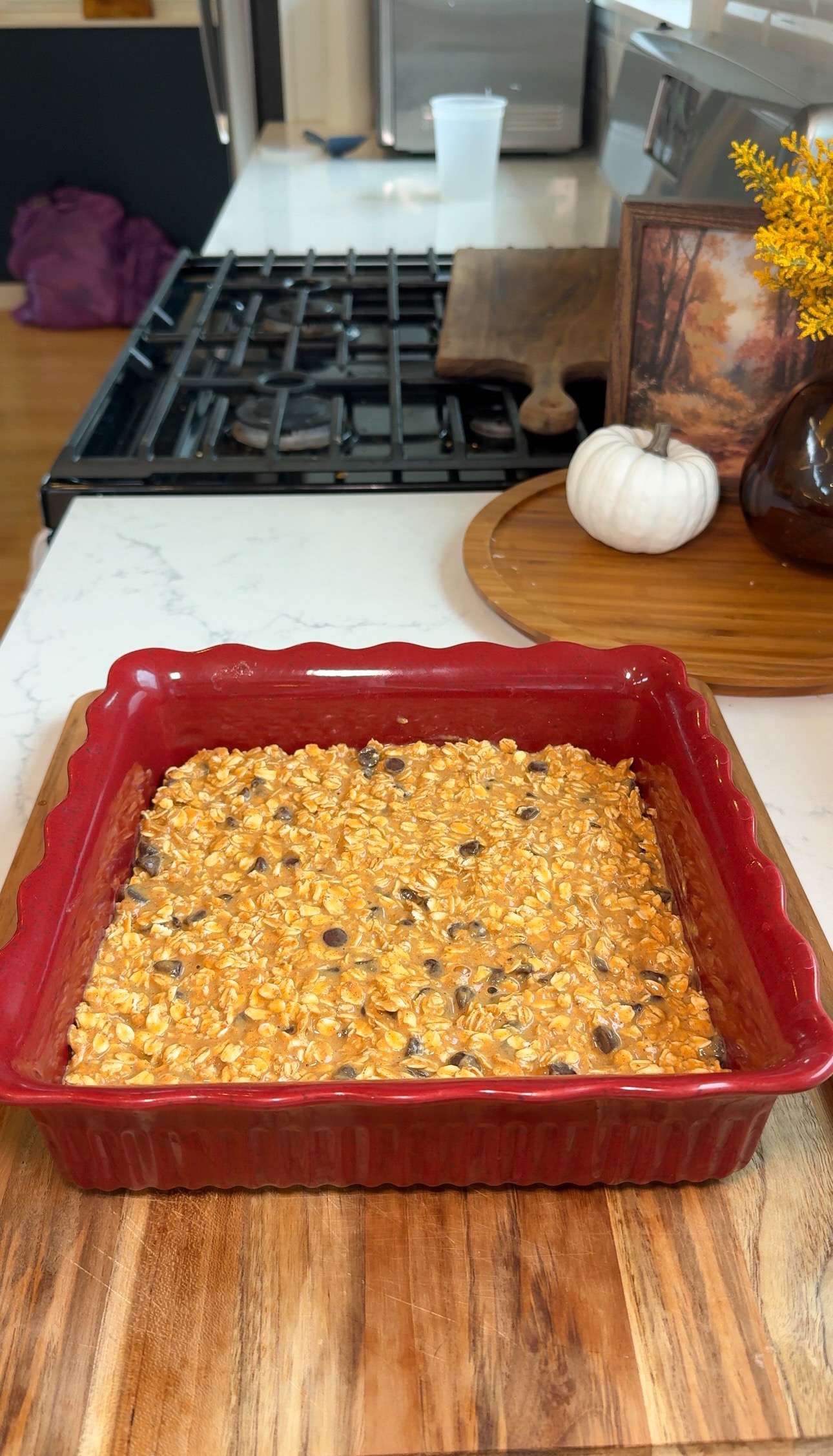 A red baking dish filled with an unbaked mixture of oats and chocolate chips sits on a kitchen counter. In the background are autumn decorations, including a white pumpkin and a vase with yellow flowers.