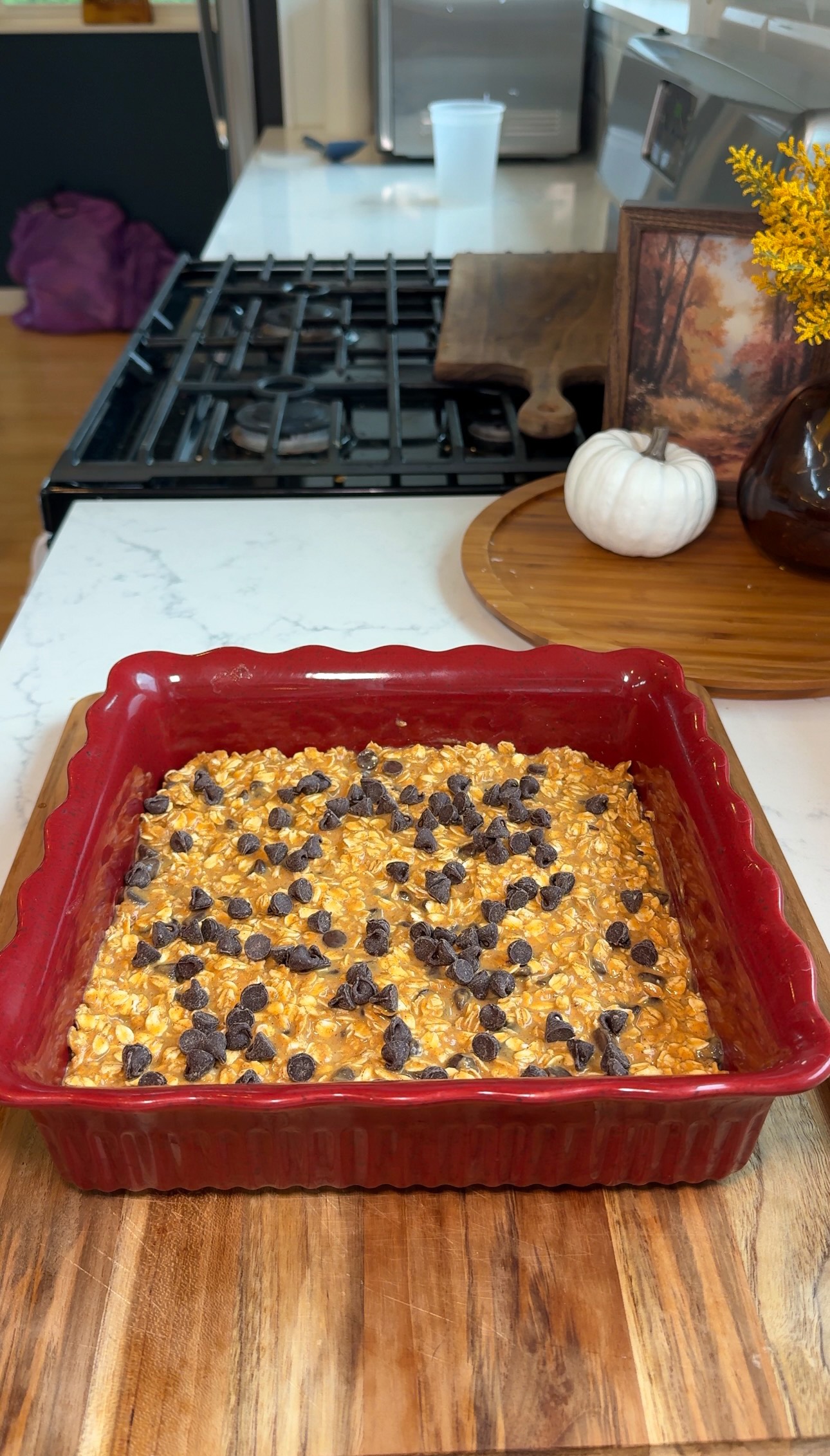 A red baking dish with oatmeal topped with chocolate chips sits on a kitchen counter. In the background are a white pumpkin, a vase with yellow flowers, and a stove.