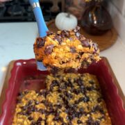 A hand holds a blue spatula lifting a slice of baked oatmeal with chocolate chips from a red baking dish. A pumpkin and yellow flowers decorate the background.