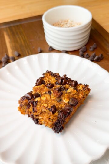 A square oatmeal chocolate chip bar sits on a white scalloped plate, with scattered chocolate chips nearby and a white bowl filled with oats in the background.