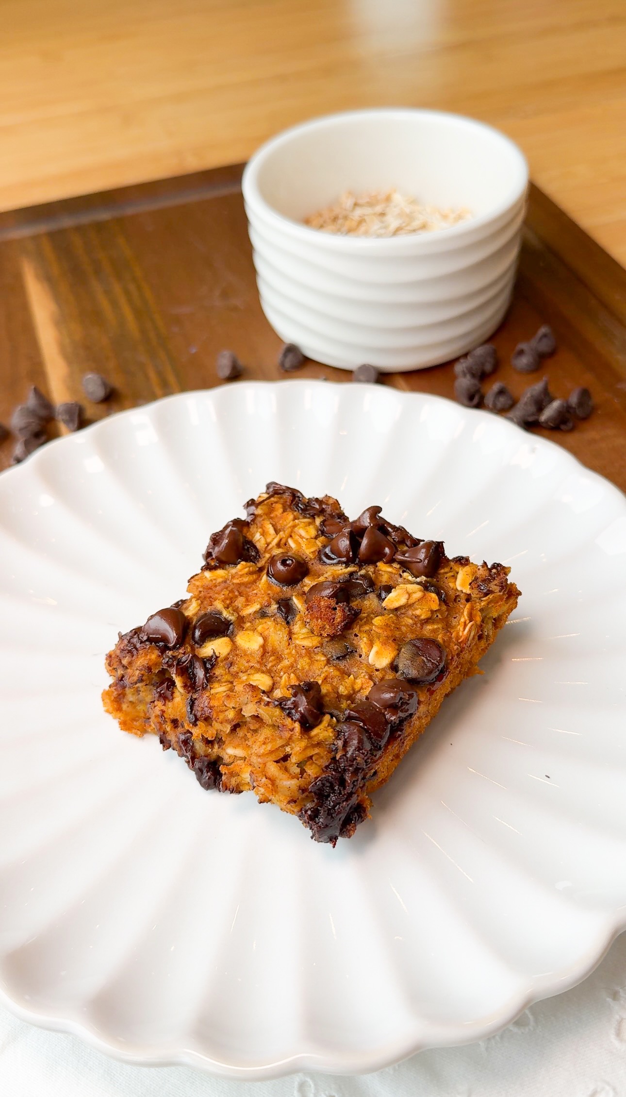 A square oatmeal chocolate chip bar sits on a white scalloped plate, with scattered chocolate chips nearby and a white bowl filled with oats in the background.