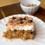 A slice of cake topped with whipped cream, chocolate chips, and crushed nuts sits on a white plate. A small bowl with extra toppings is in the background on a wooden surface.