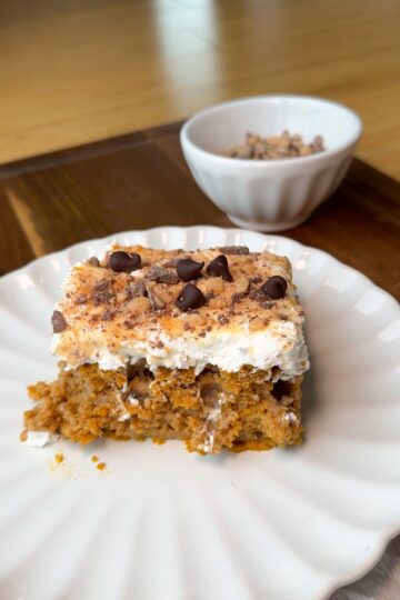 A slice of cake topped with whipped cream, chocolate chips, and crushed nuts sits on a white plate. A small bowl with extra toppings is in the background on a wooden surface.