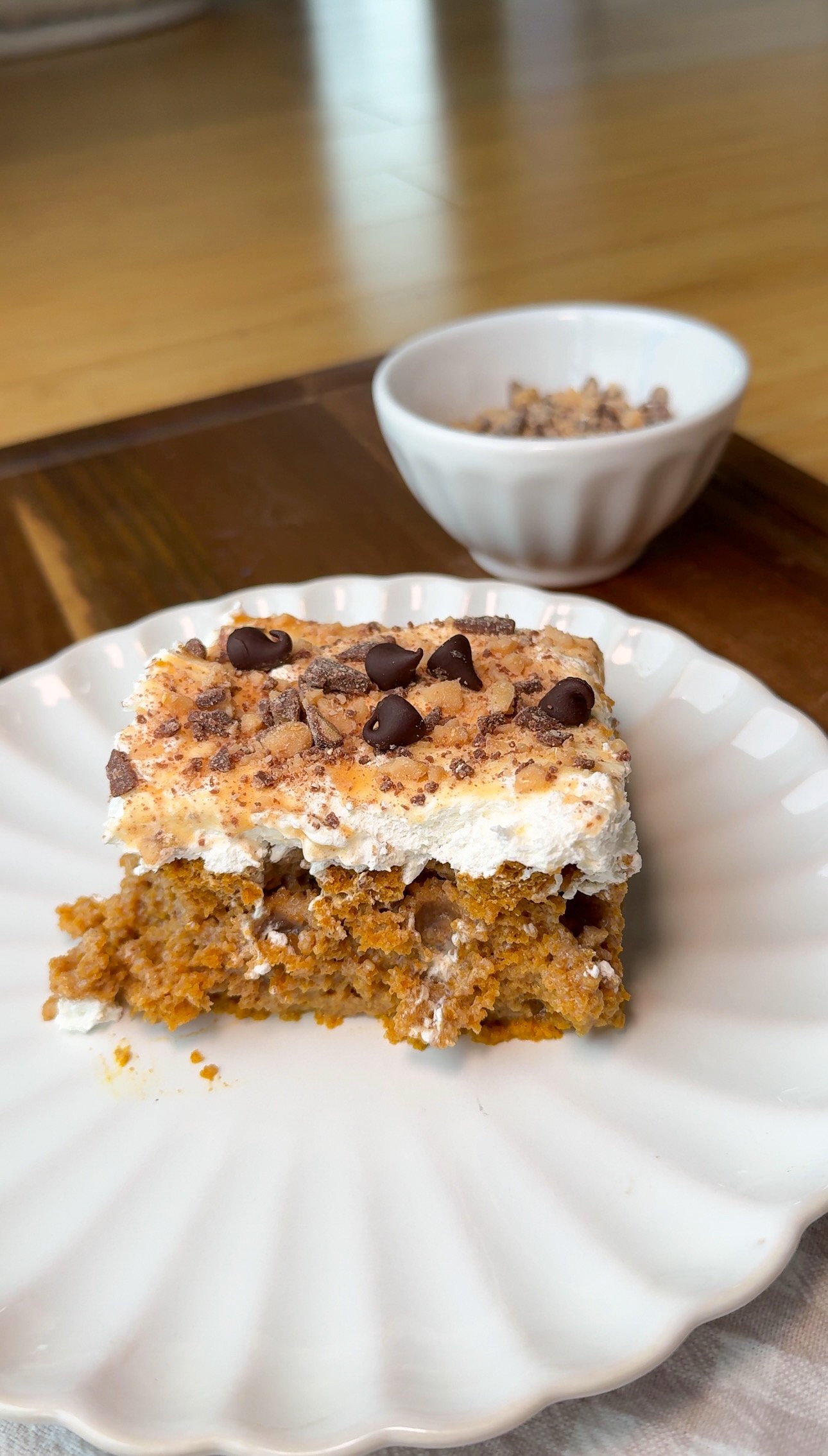 A slice of cake topped with whipped cream, chocolate chips, and crushed nuts sits on a white plate. A small bowl with extra toppings is in the background on a wooden surface.