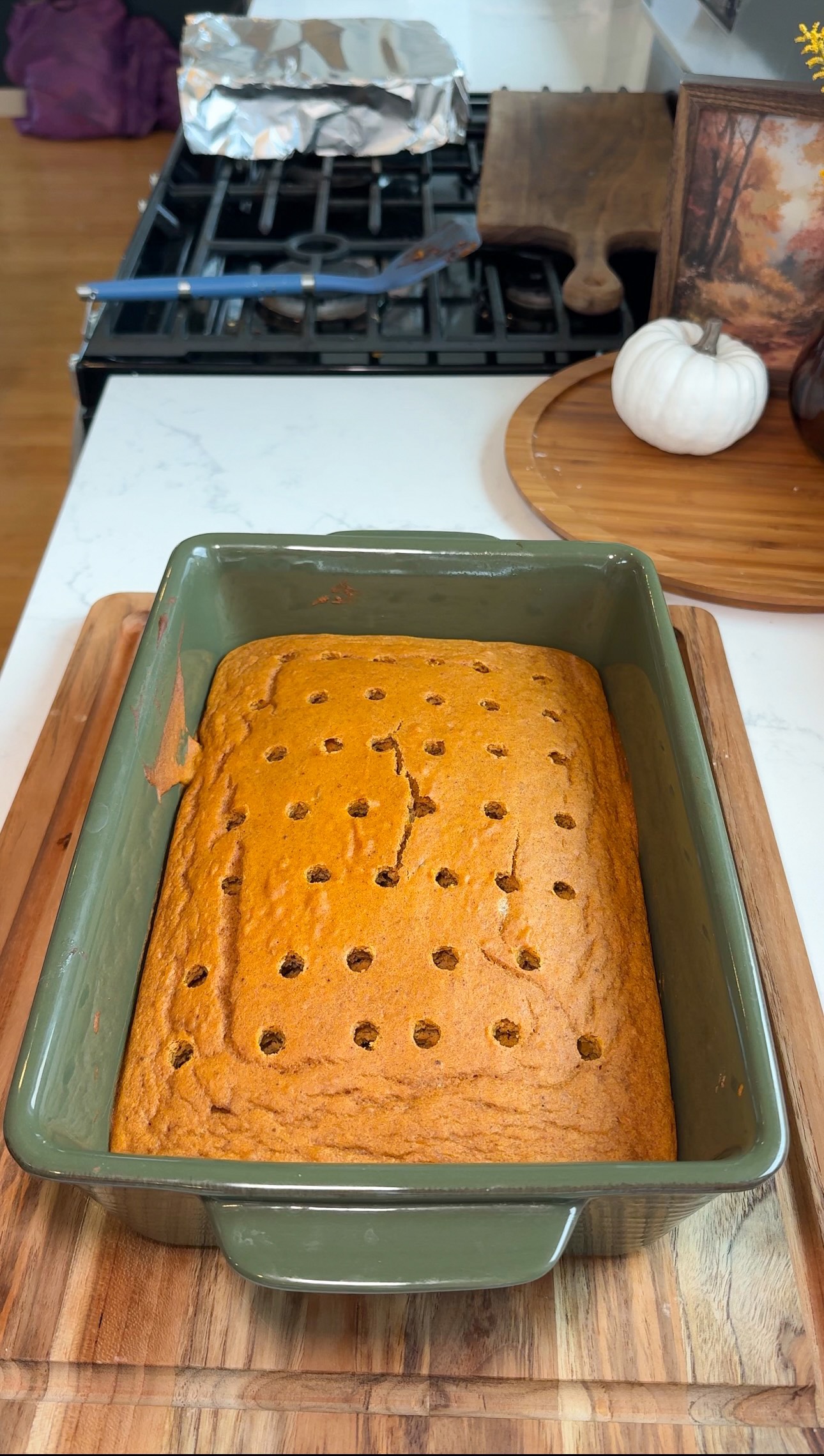 A rectangular pumpkin cake in a green baking dish sits on a wooden board. The cake has evenly spaced holes on top, and kitchen items are visible in the background.
